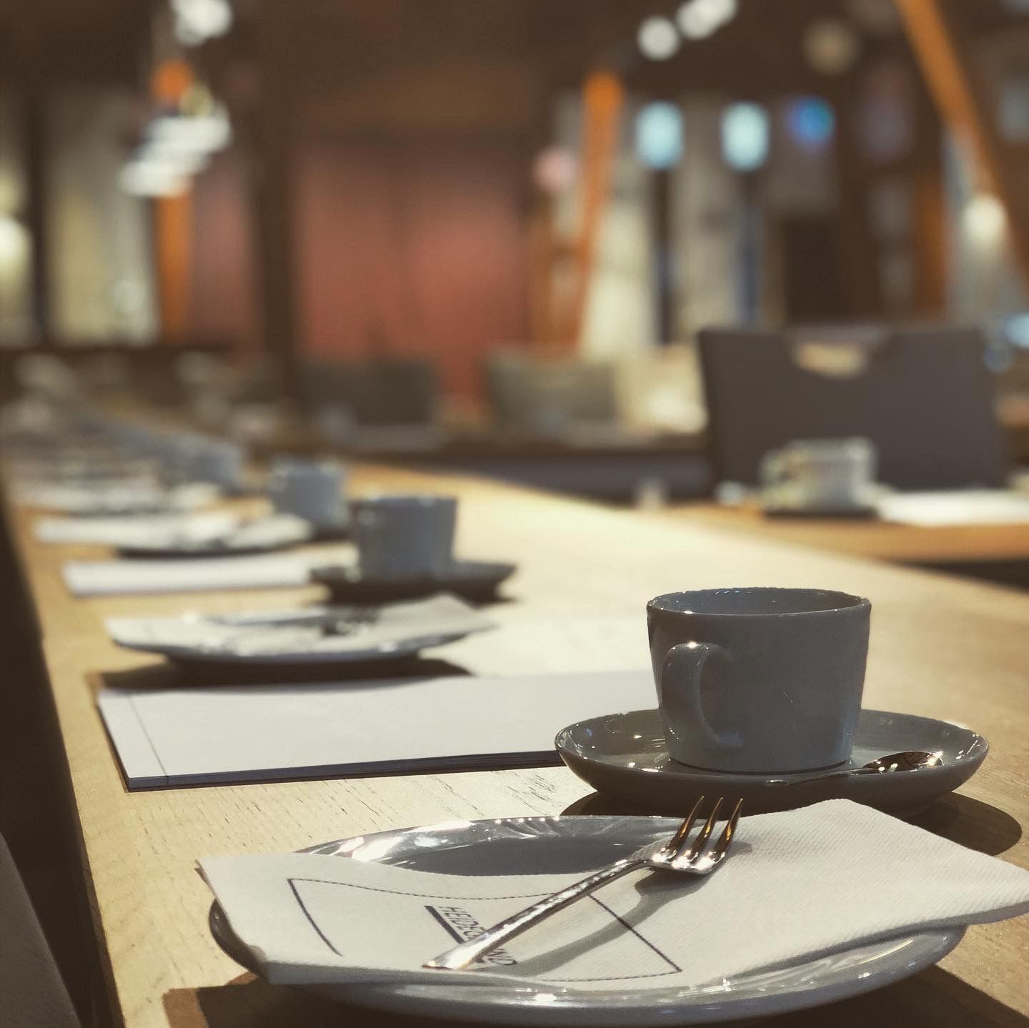 In einem sanft beleuchteten Restaurant erstreckt sich ein langer Tisch mit weißen Tassen, Untertassen, Servietten und Gabeln bis in die Ferne.