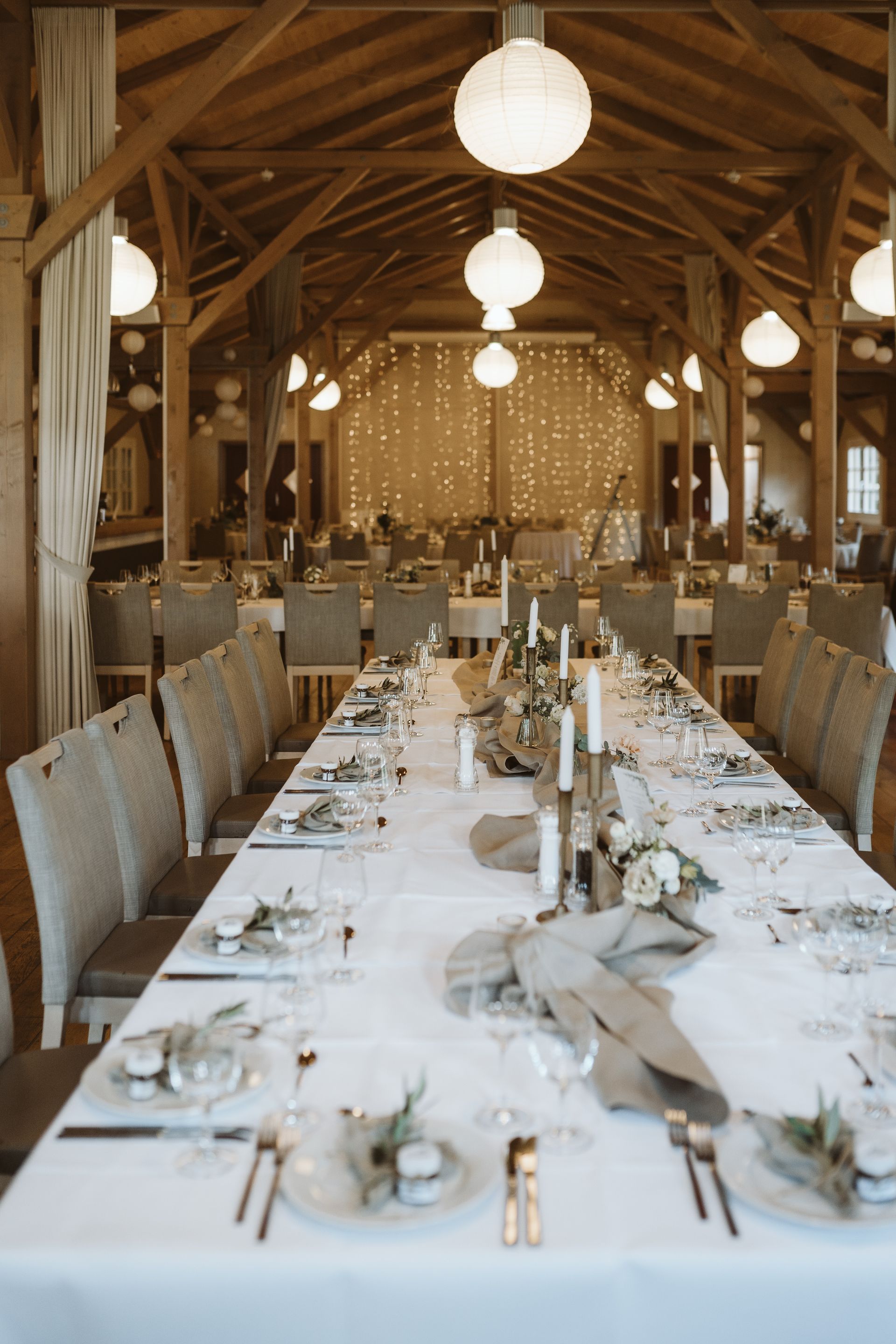 Eine lange Tafel, gedeckt für eine Hochzeit in einer rustikalen Holzscheune mit weißen Papierlaternen und Lichterketten.