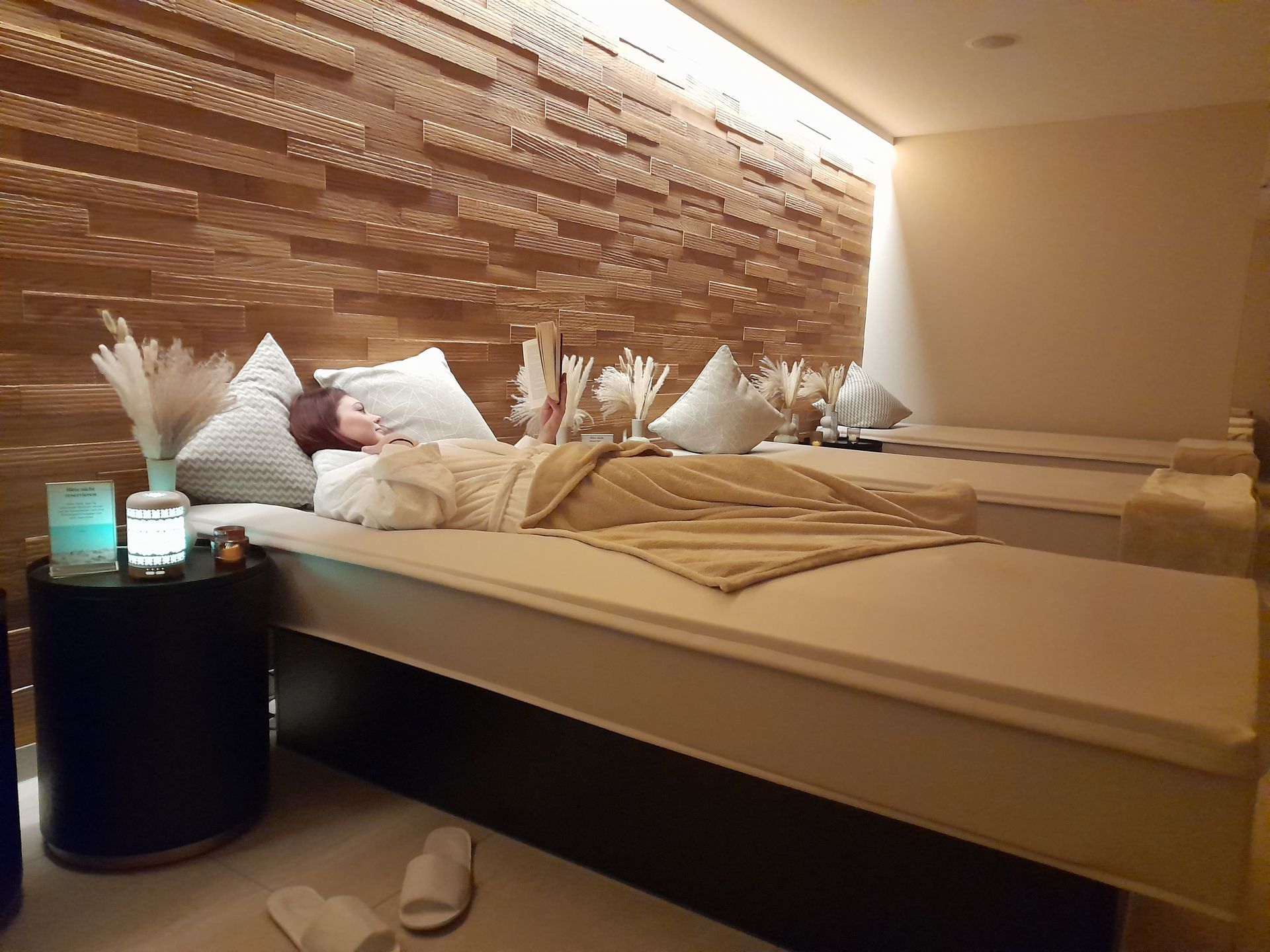 Eine Person entspannt sich auf einer Massageliege in einem Spa-Raum mit strukturierten Holzwänden und gedämpftem, stimmungsvollem Licht.