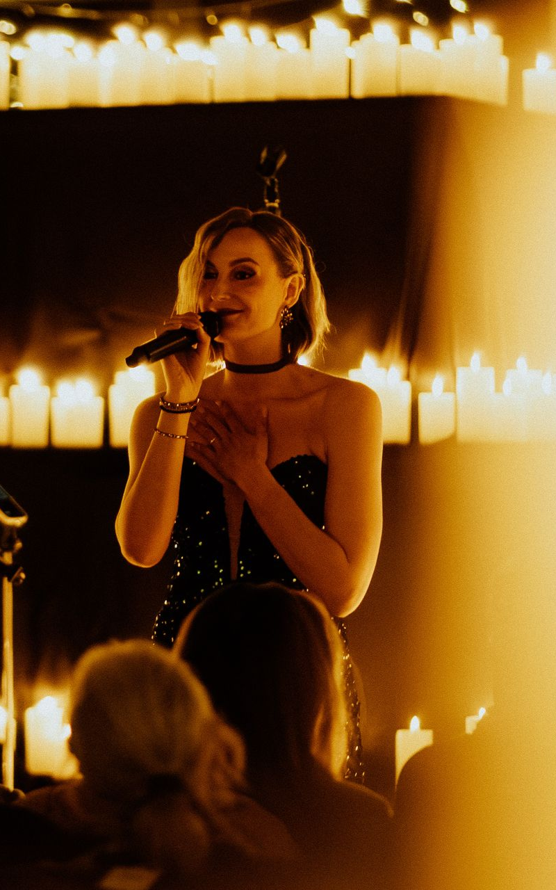 A singer in a dark, sequined dress appears on stage against a backdrop of numerous glowing white candles.