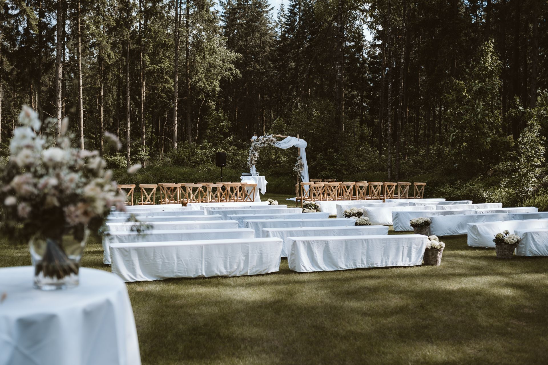Eine im Freien stattfindende Hochzeitszeremonie in einem Wald mit Reihen weiß gedeckter Bänke, die auf einen Blumenbogen auf einer Rasenfläche ausgerichtet sind.
