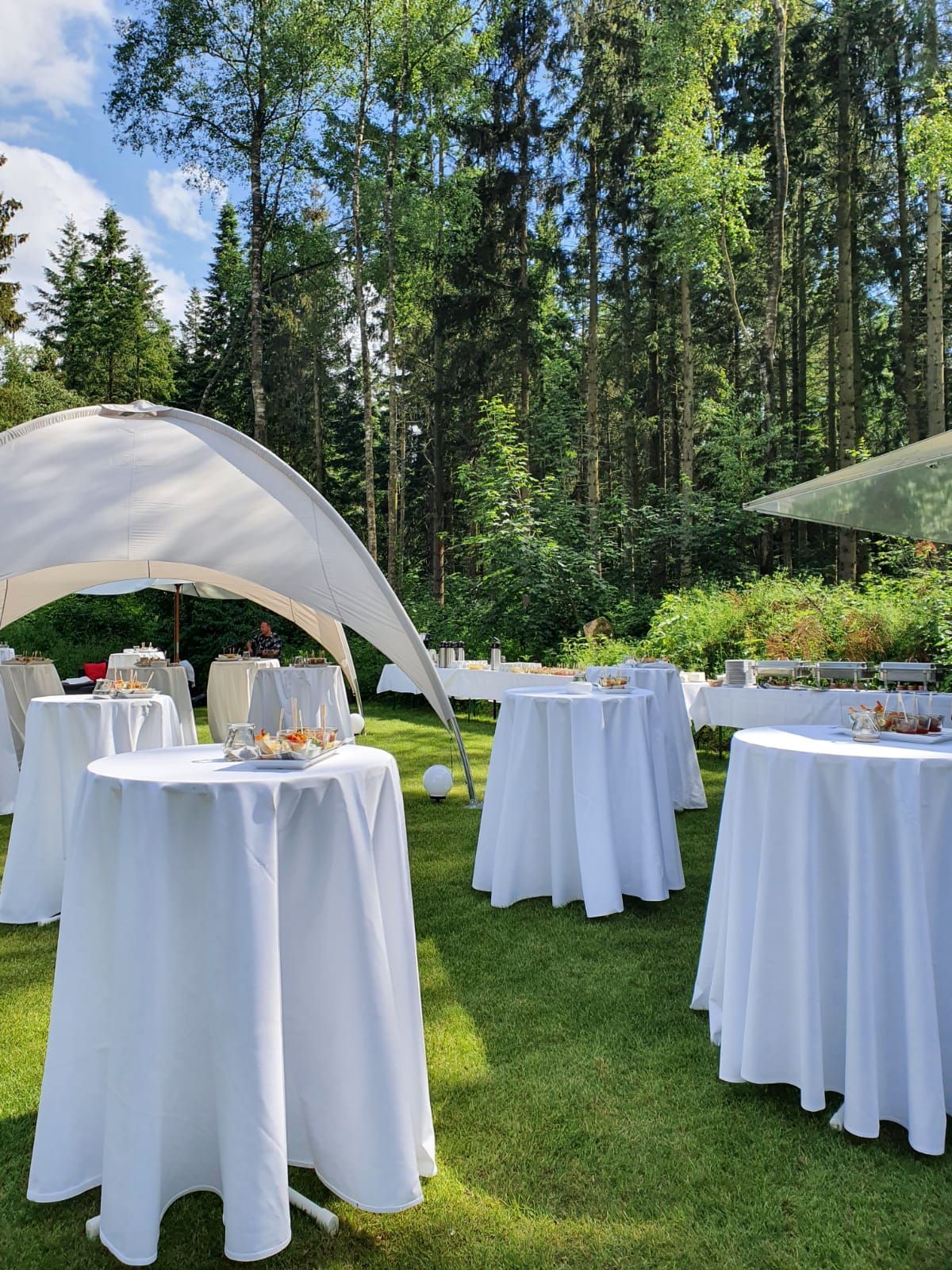 Weiße Stehtische mit Tischdecken stehen auf einer Rasenfläche unter Bäumen, darüber ein weißes Sonnensegel.