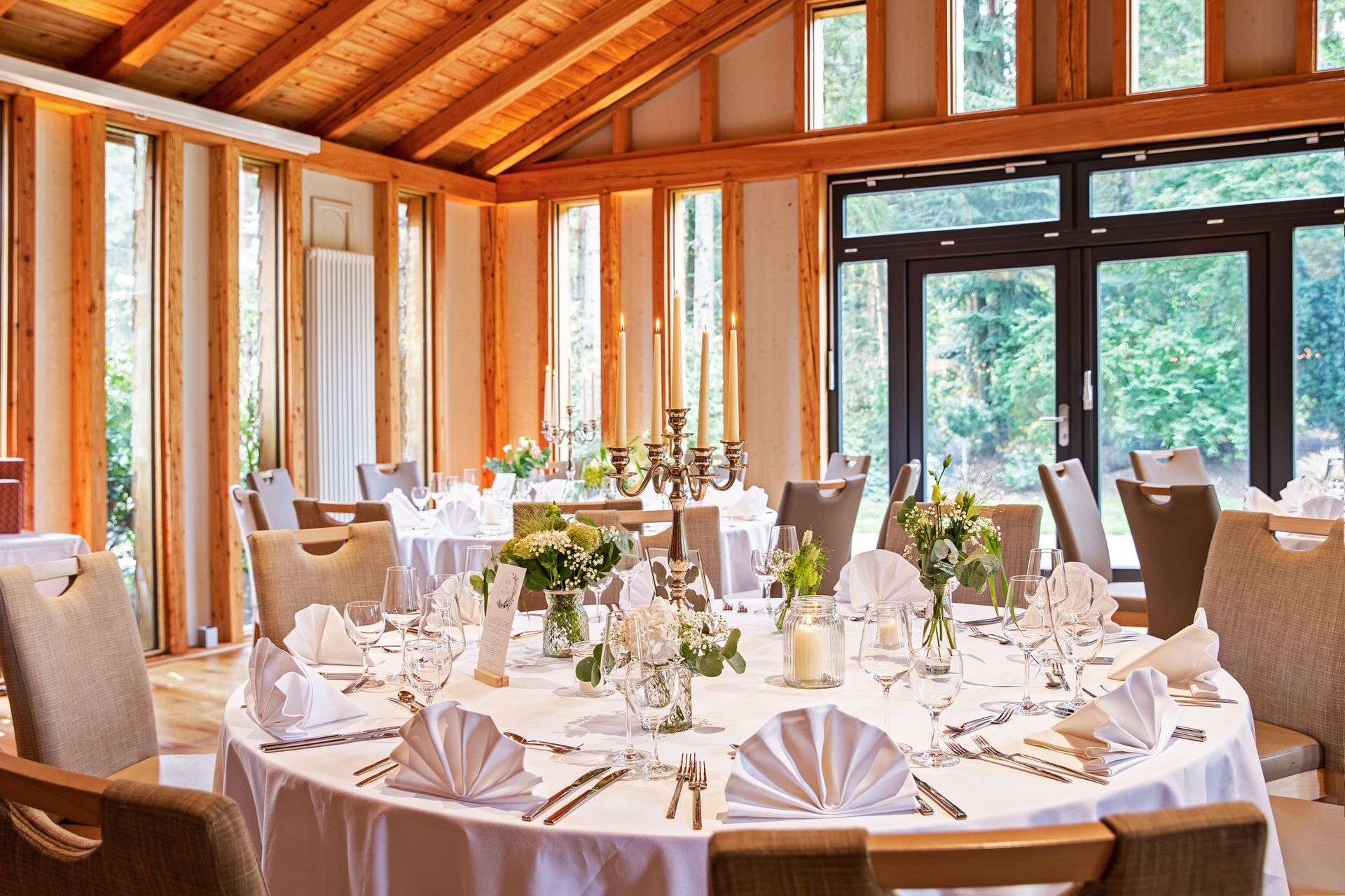 A bright, sun-drenched dining room set for a formal event with round tables, white tablecloths, folded napkins, and exposed wooden beams.