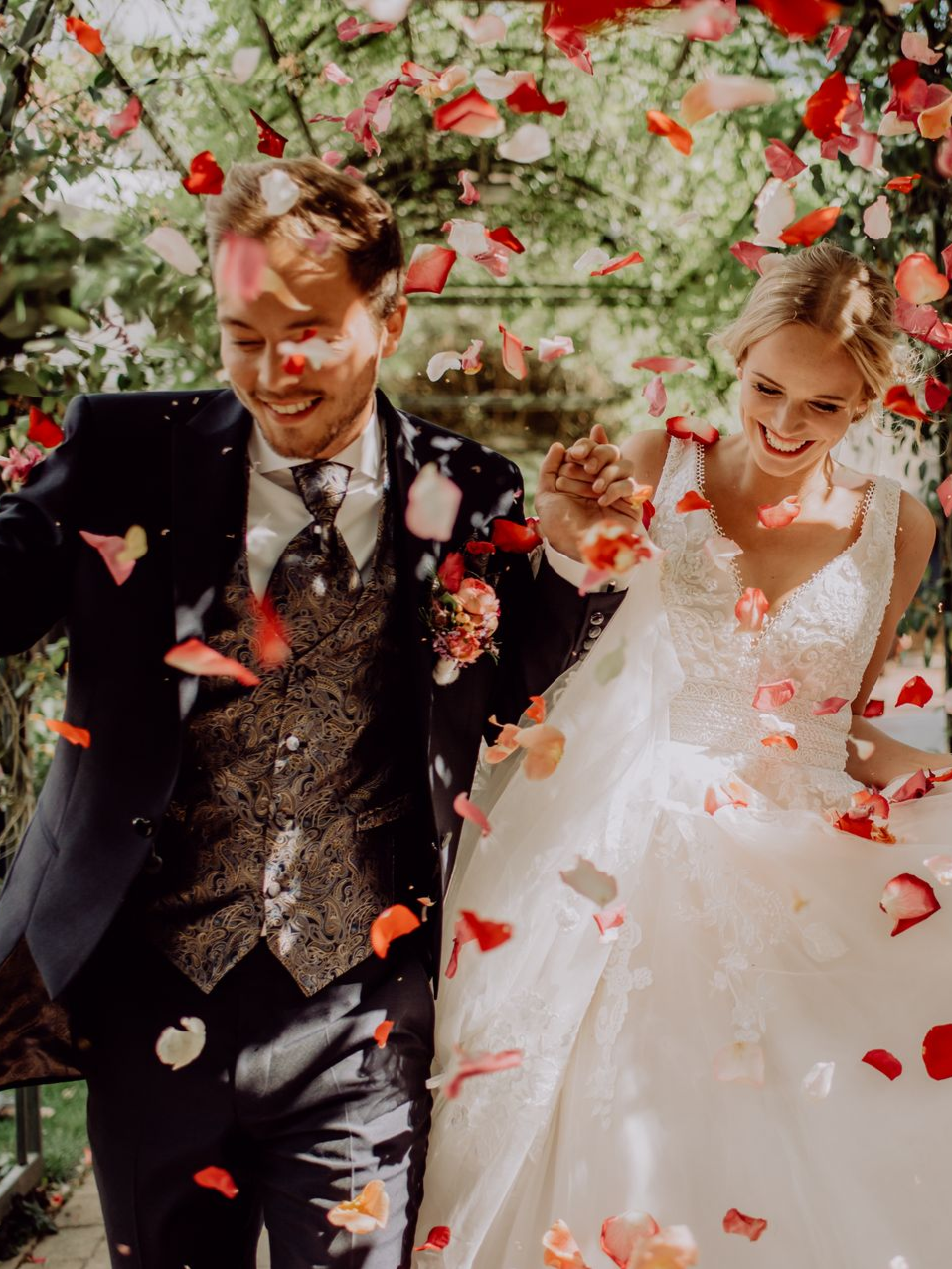 Drei Personen in formeller Kleidung sitzen auf dem Rücksitz eines Cabrios, halten Champagnergläser in der Hand und lächeln inmitten von Grün.