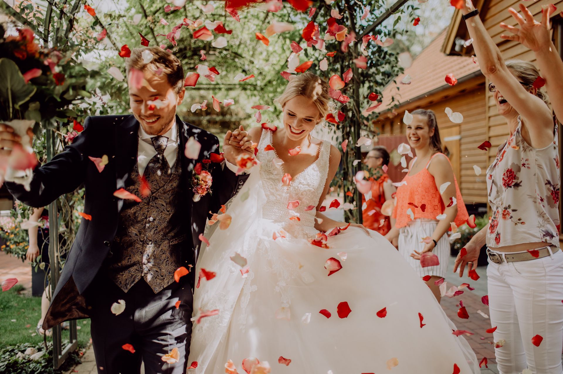 A bride and groom walk through a floral arch while guests scatter rose petals and celebrate their wedding in an outdoor garden.