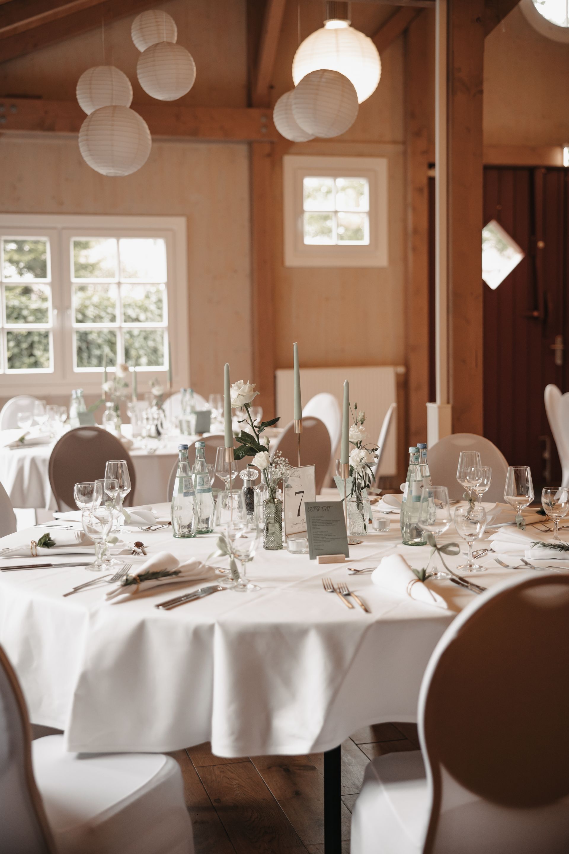 Runde Esstische, gedeckt für eine Hochzeit in einem rustikalen, holzgetäfelten Raum mit weißer Papierlaternenbeleuchtung.