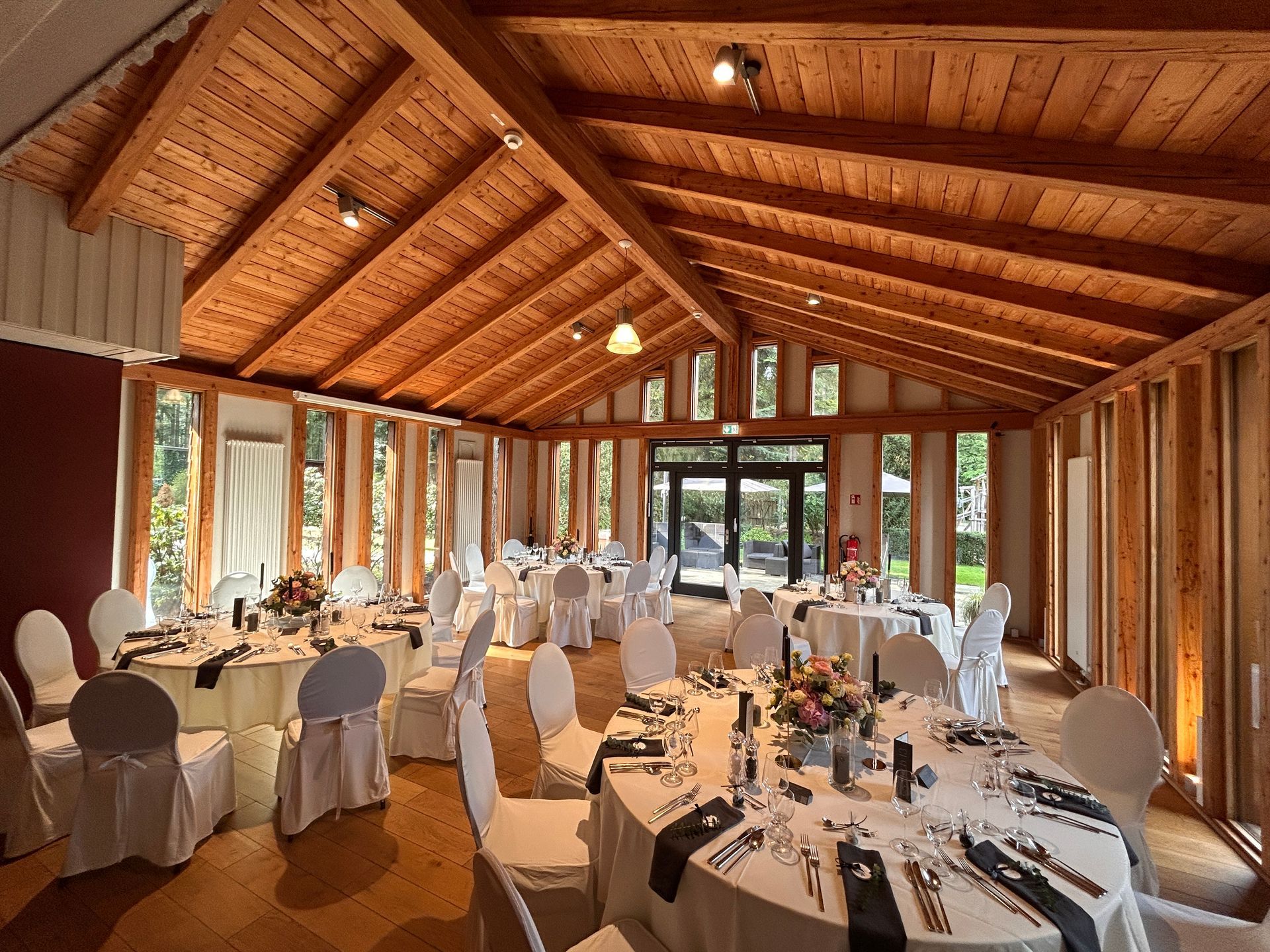 Ein Speisesaal mit gewölbter Holzdecke, runden Tischen, die für eine Veranstaltung gedeckt sind, und großen Fenstern mit Blick ins Grüne.