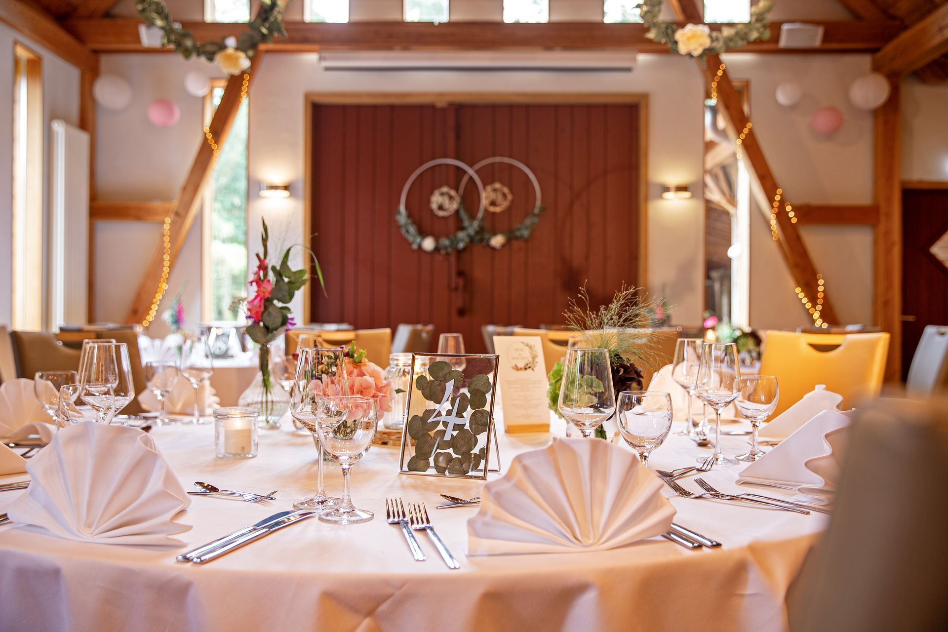 A round table set for a wedding reception in a barn, with white tablecloths, floral arrangements, and place cards.