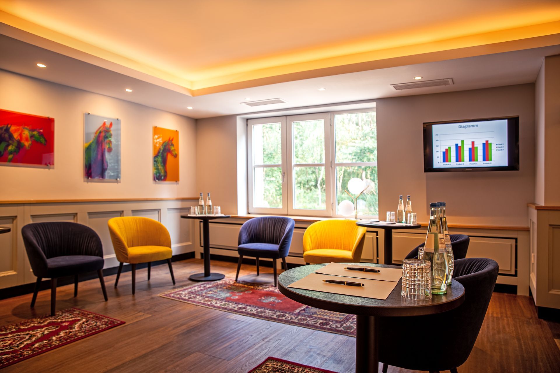 Ein Loungebereich mit blauen und gelben Stühlen um kleine Tische, farbenfroher Wandkunst und einem digitalen Diagramm an der Wand.