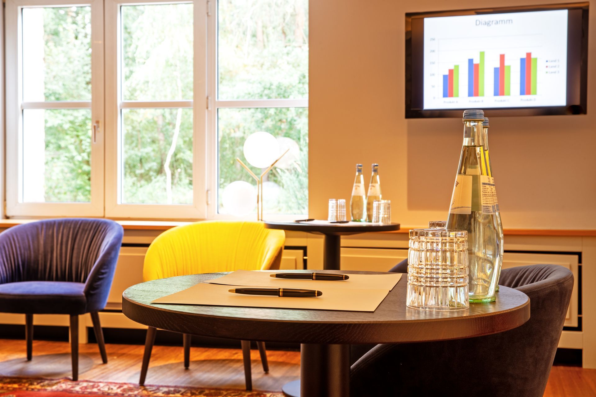 Ein Besprechungsraum mit einem gelben Stuhl, einem violetten Stuhl, einem runden Tisch mit Stiften und Wasser sowie einem Bildschirm, auf dem ein Balkendiagramm angezeigt wird.