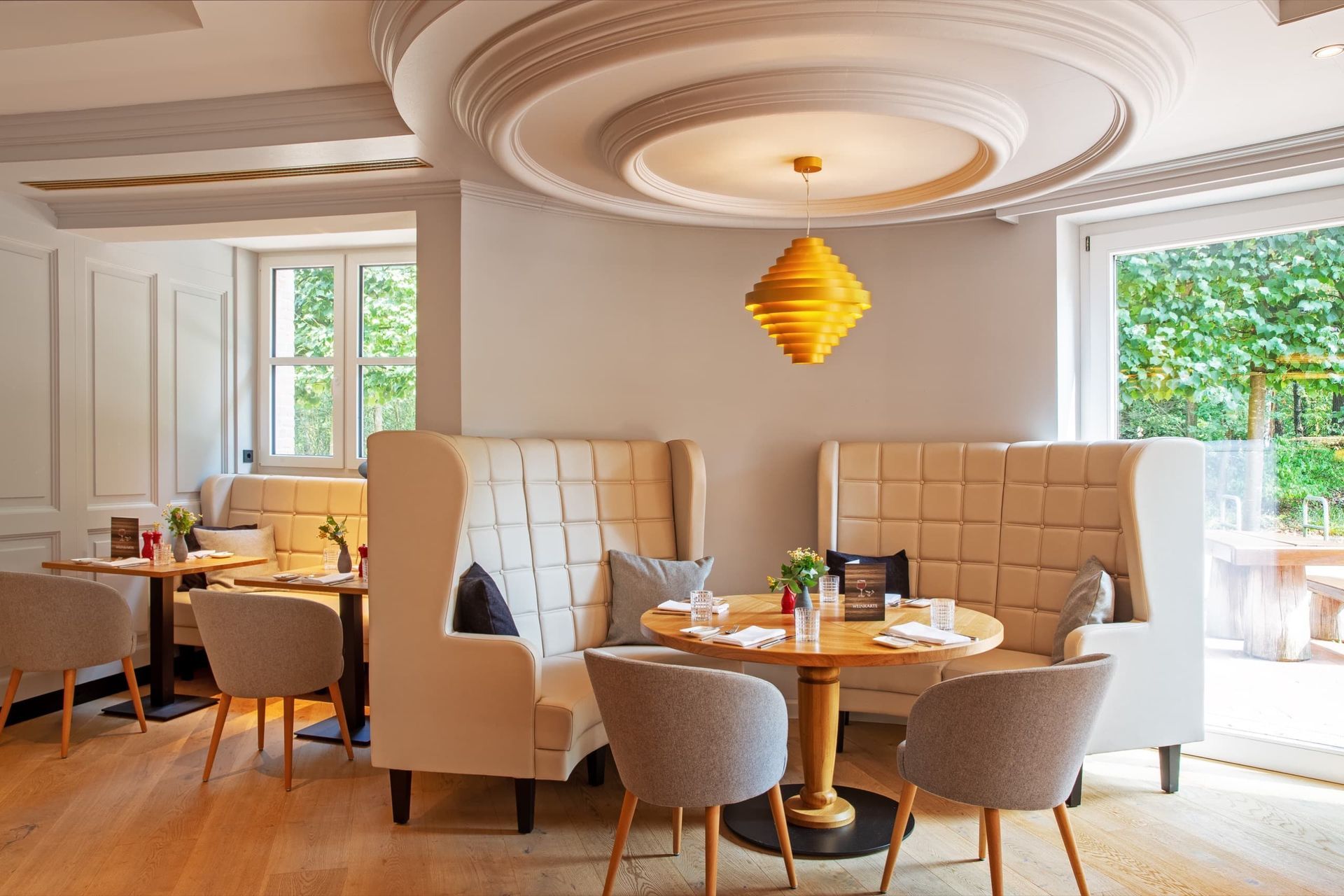 A bright, elegant restaurant interior featuring beige-colored seating alcoves with high backs, round wooden tables, gray chairs, and a yellow lamp.