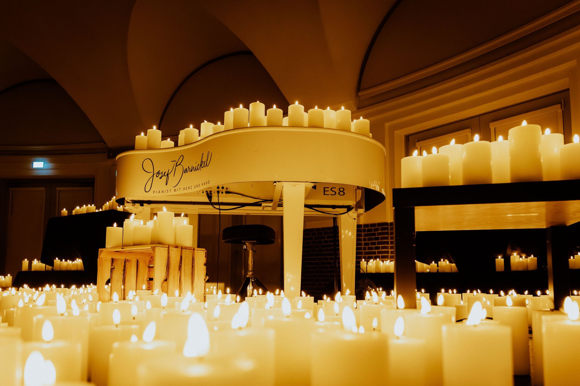 Eine Person singt vor Publikum in ein Mikrofon, umgeben von vielen brennenden Kerzen in einem warmen, schwach beleuchteten Raum.