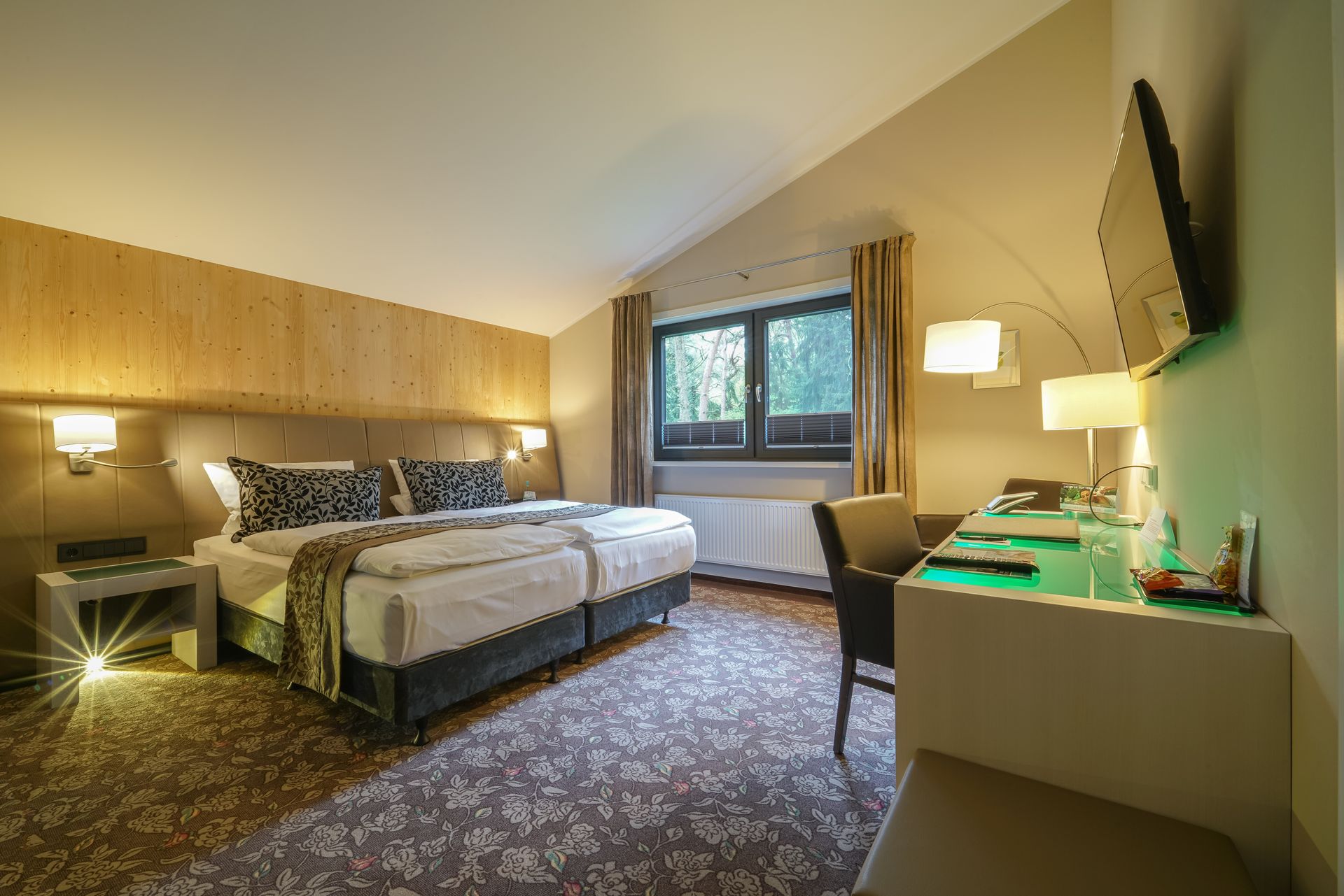 A modern hotel room with a neatly made double bed, dark patterned pillows, wall sconces, and a small sitting area.
