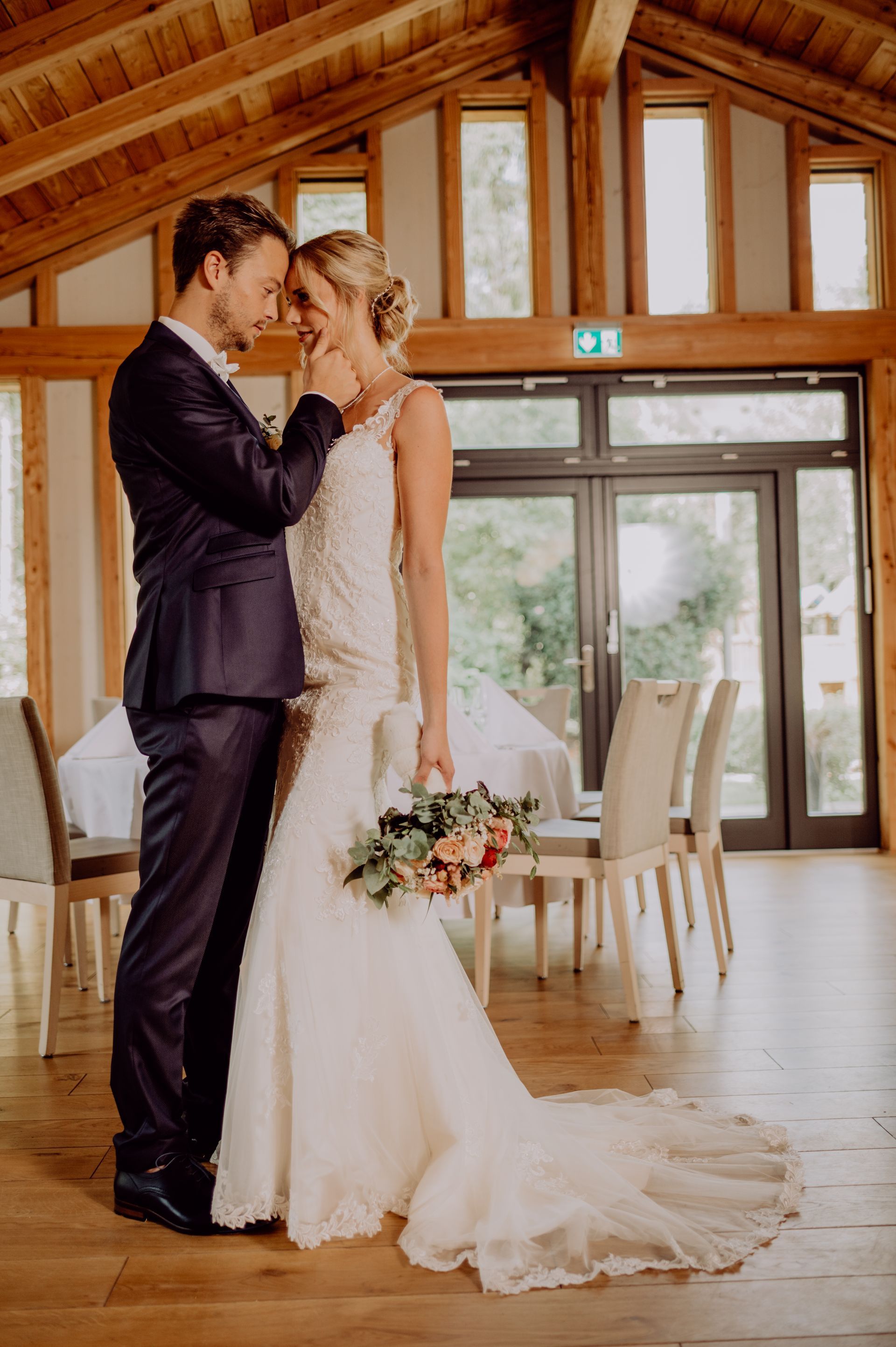 Ein Paar in Hochzeitskleidung genießt einen intimen Moment, einander gegenüber in einer rustikalen Halle mit Holzbalken.