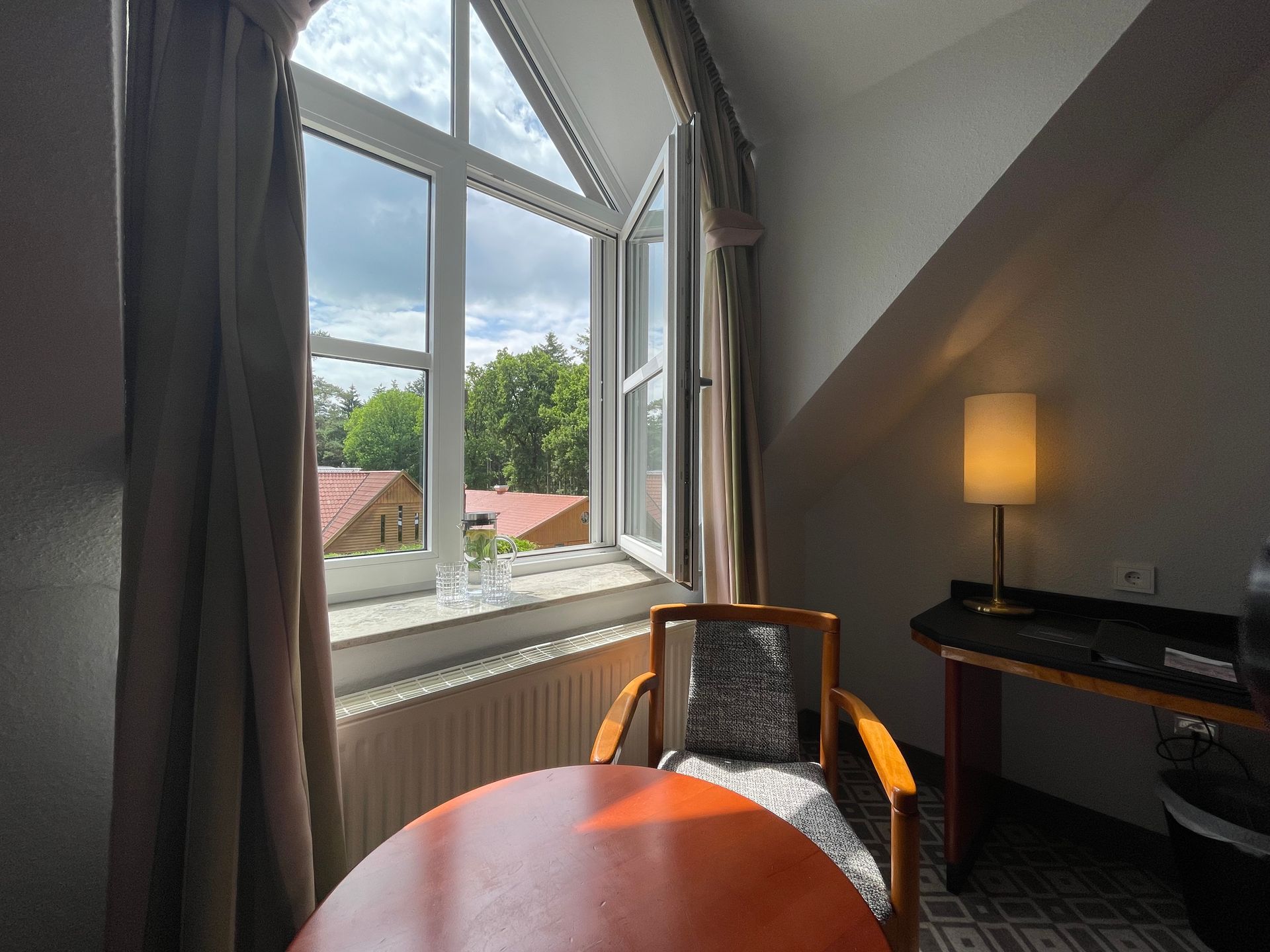 Ein gemütliches Zimmer mit rundem Tisch, Holzstuhl, Schreibtisch mit Lampe und einem großen Fenster mit Blick auf Bäume und Häuser.