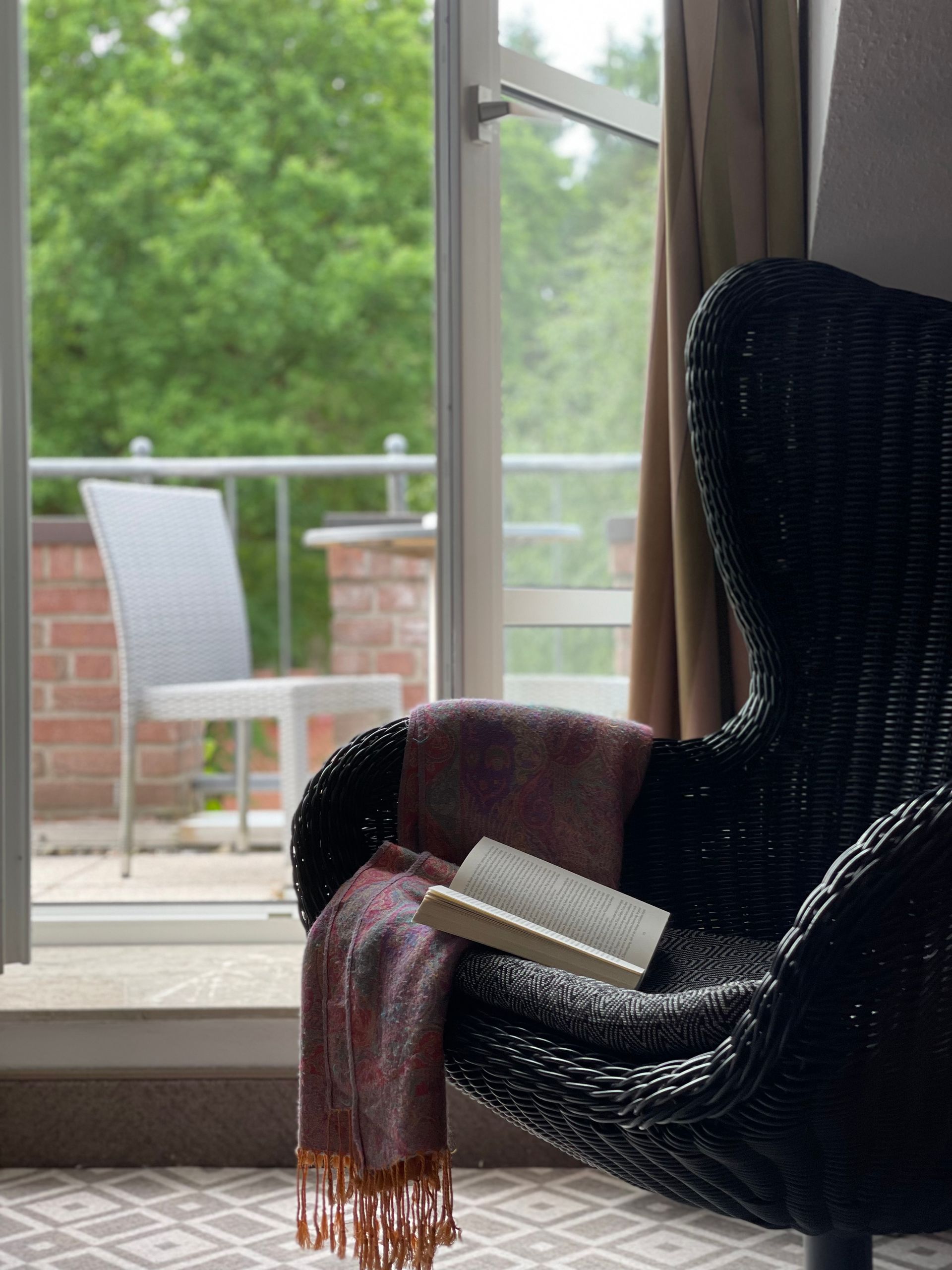Ein schwarzer Korbsessel, auf dem ein Buch und ein drapierter Schal liegen, steht an einem Fenster mit Blick auf einen Balkon und grüne Bäume.