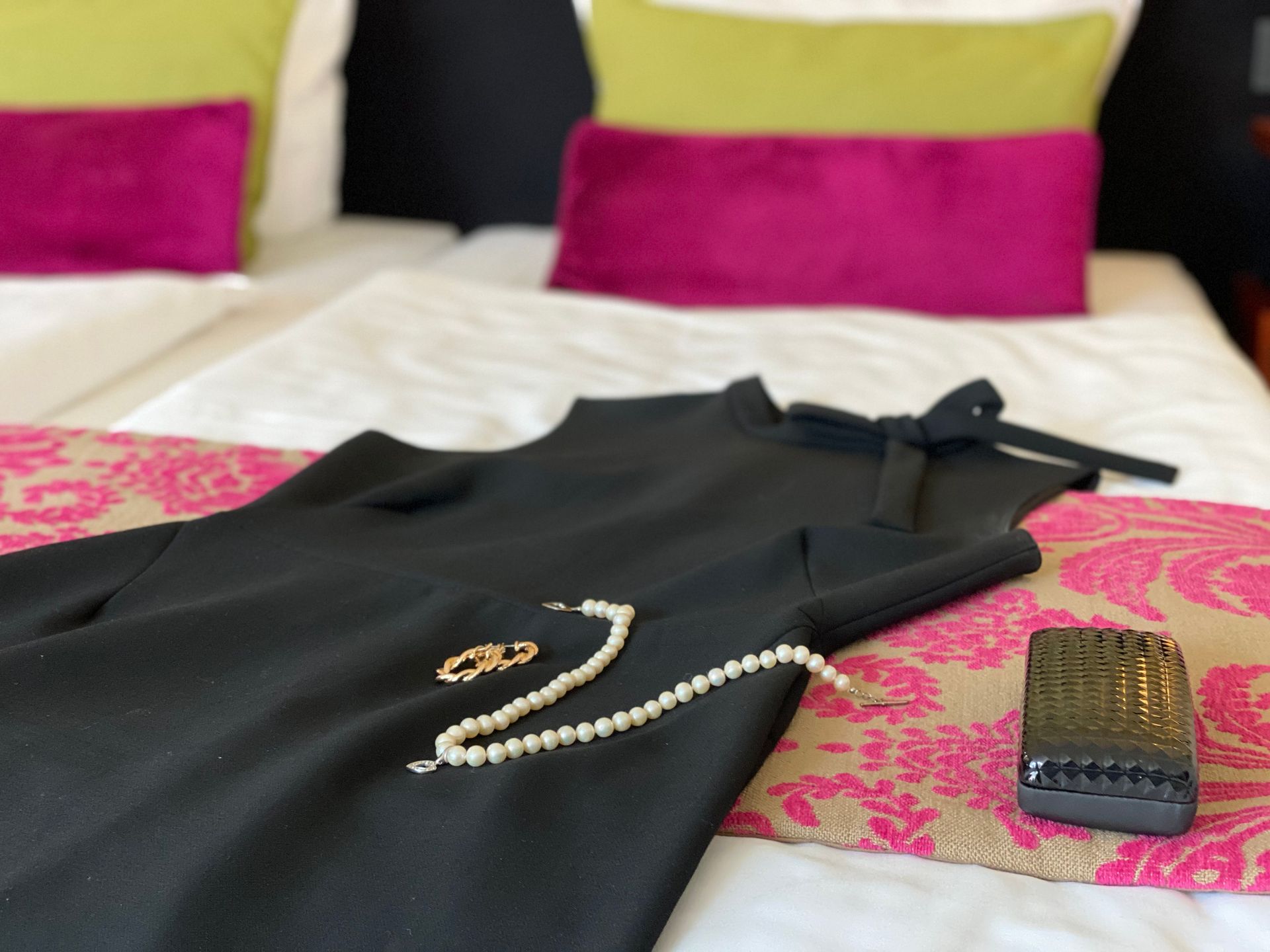 A black dress, a pearl necklace, a gold ring, and a black clutch lie on a bed covered with colorful decorative pillows.