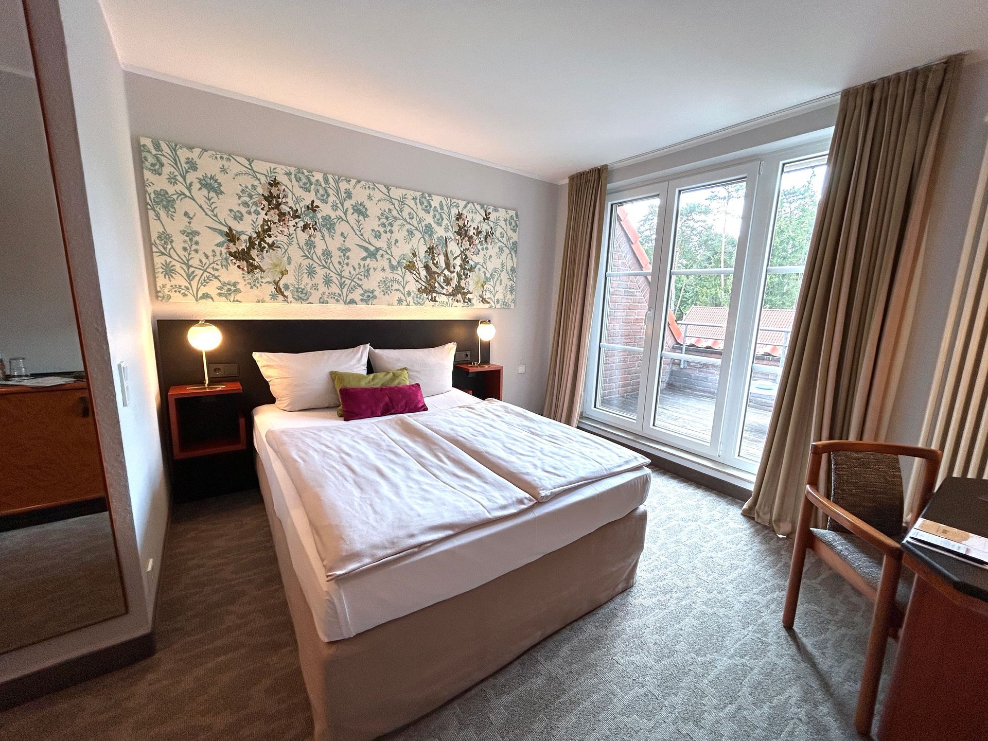 Ein gemütliches Zimmer mit rundem Tisch, Holzstuhl, Schreibtisch mit Lampe und einem großen Fenster mit Blick auf Bäume und Häuser.