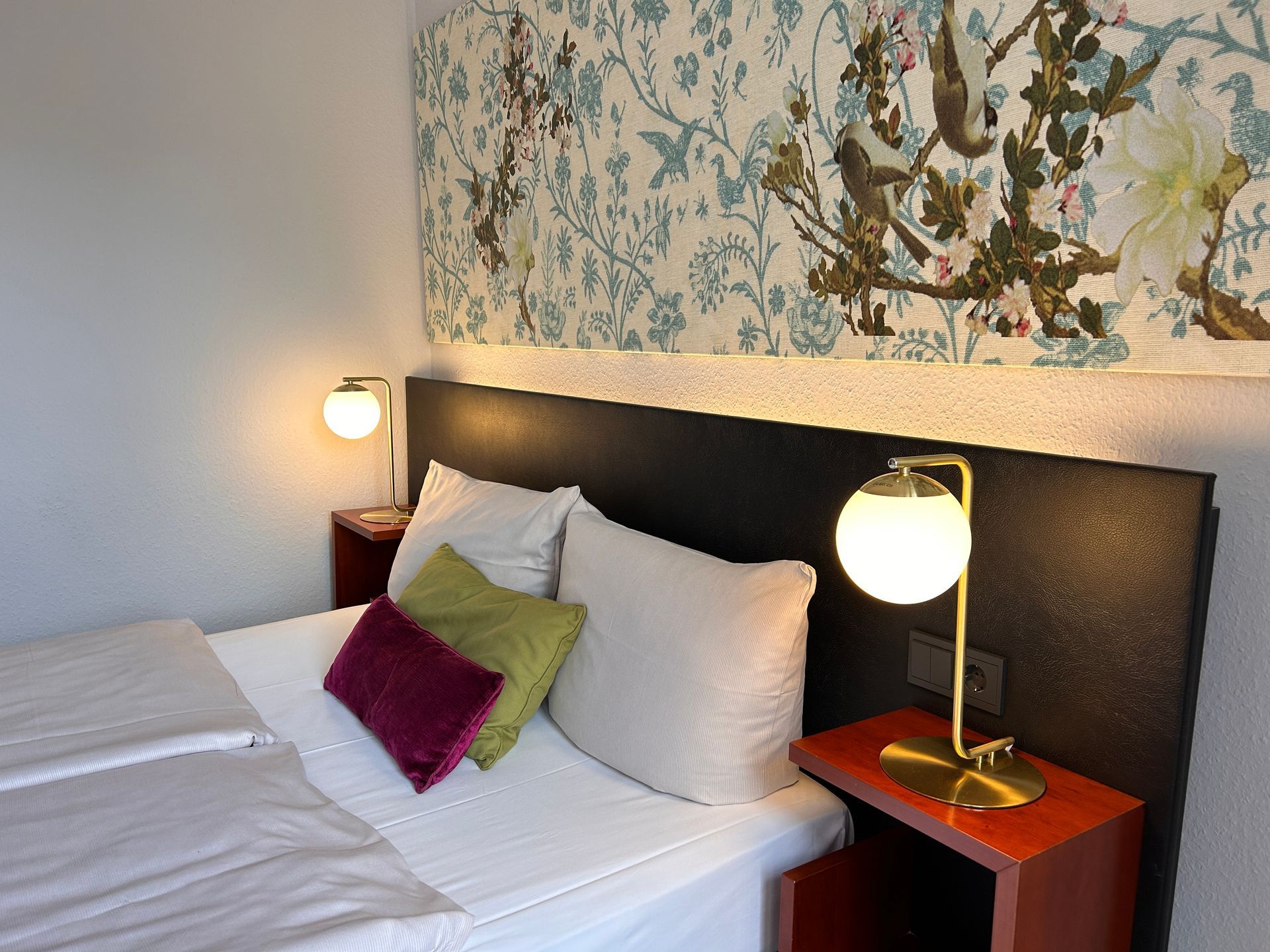 Ein modernes Hotelzimmer mit einem ordentlich gemachten Doppelbett, dunkel gemusterten Kissen, Wandlampen und einer kleinen Sitzecke.