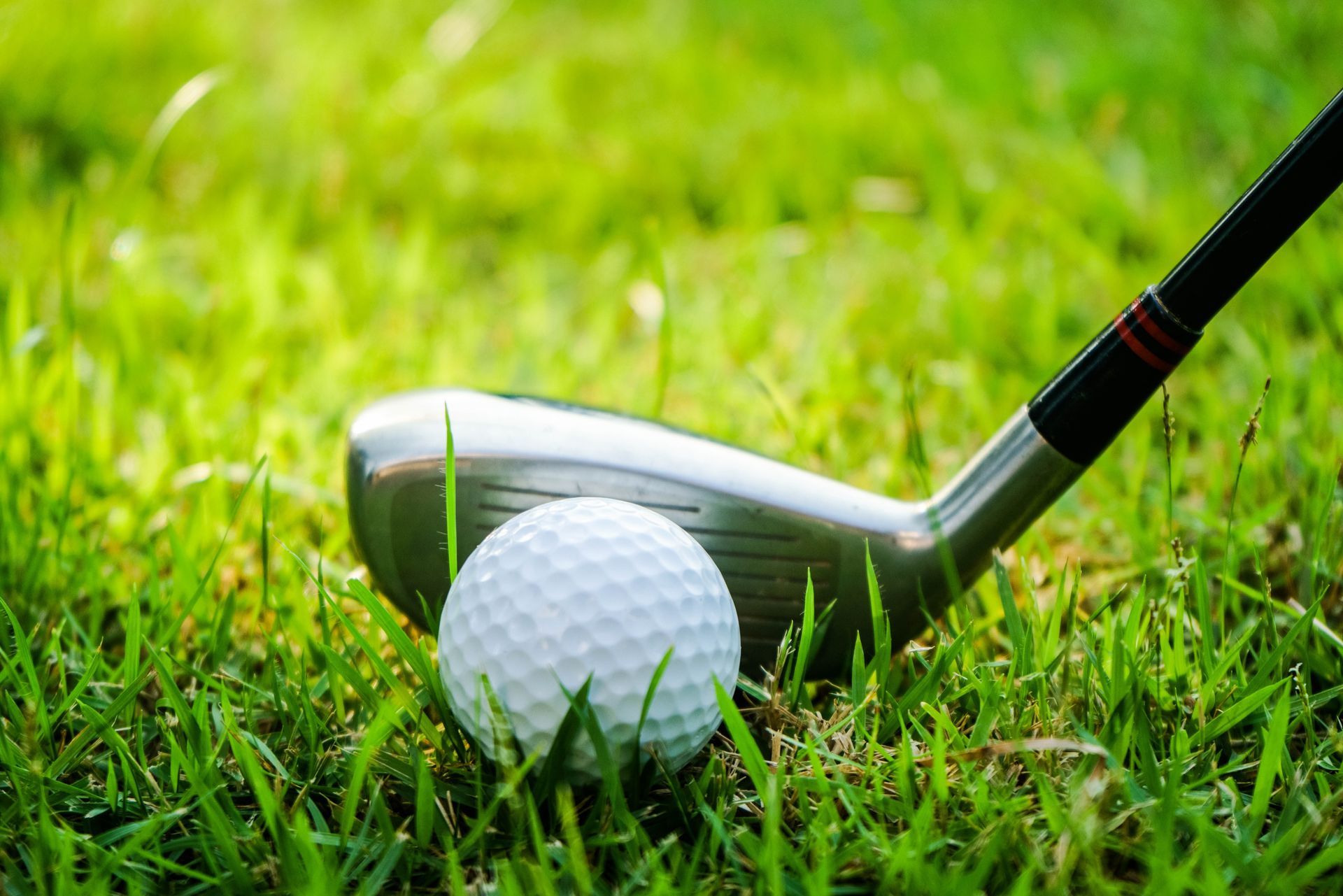 A golf ball lies on lush green grass in front of the metal head of a golf club.