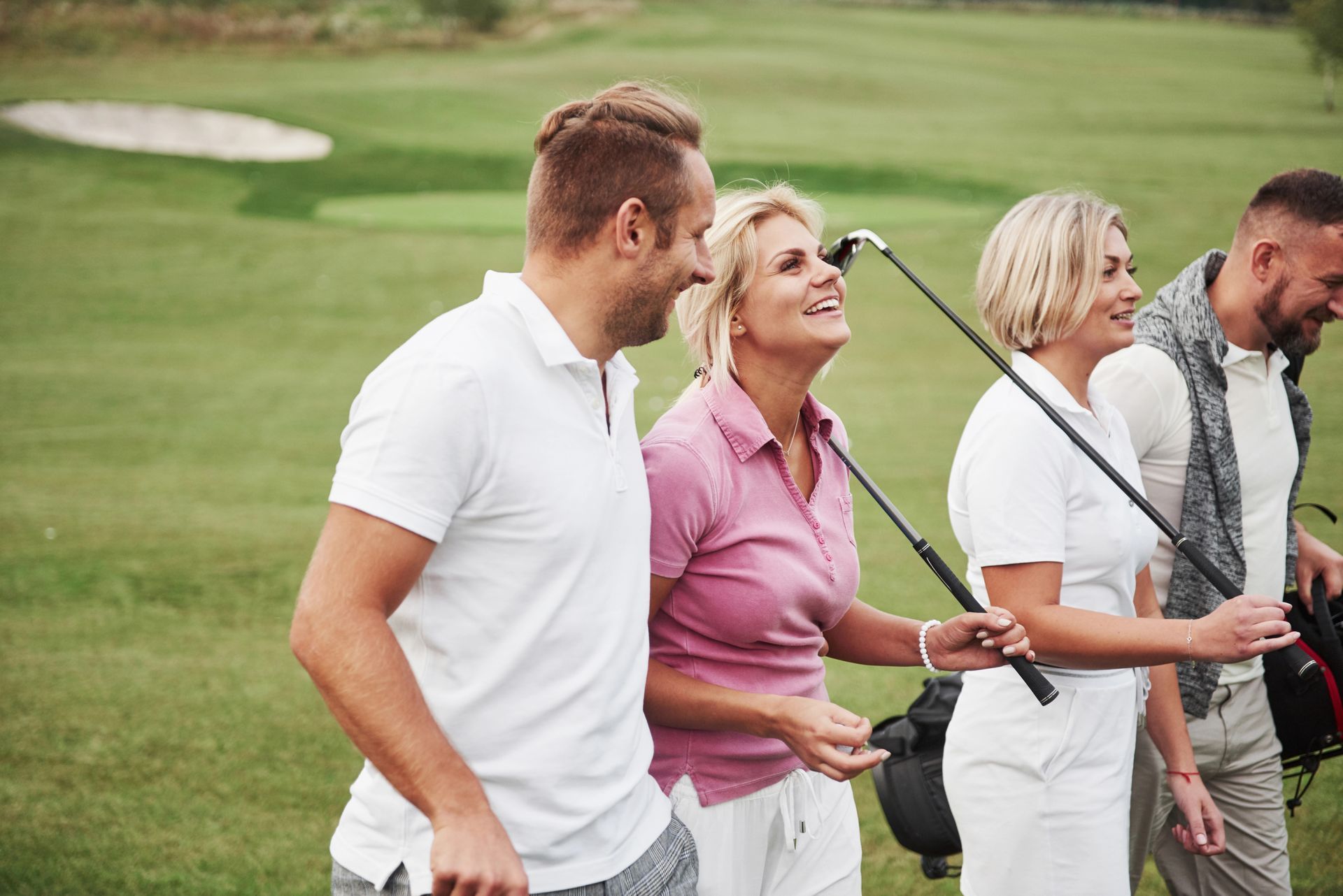 Four friends are walking across a golf course, laughing and carrying their golf clubs.