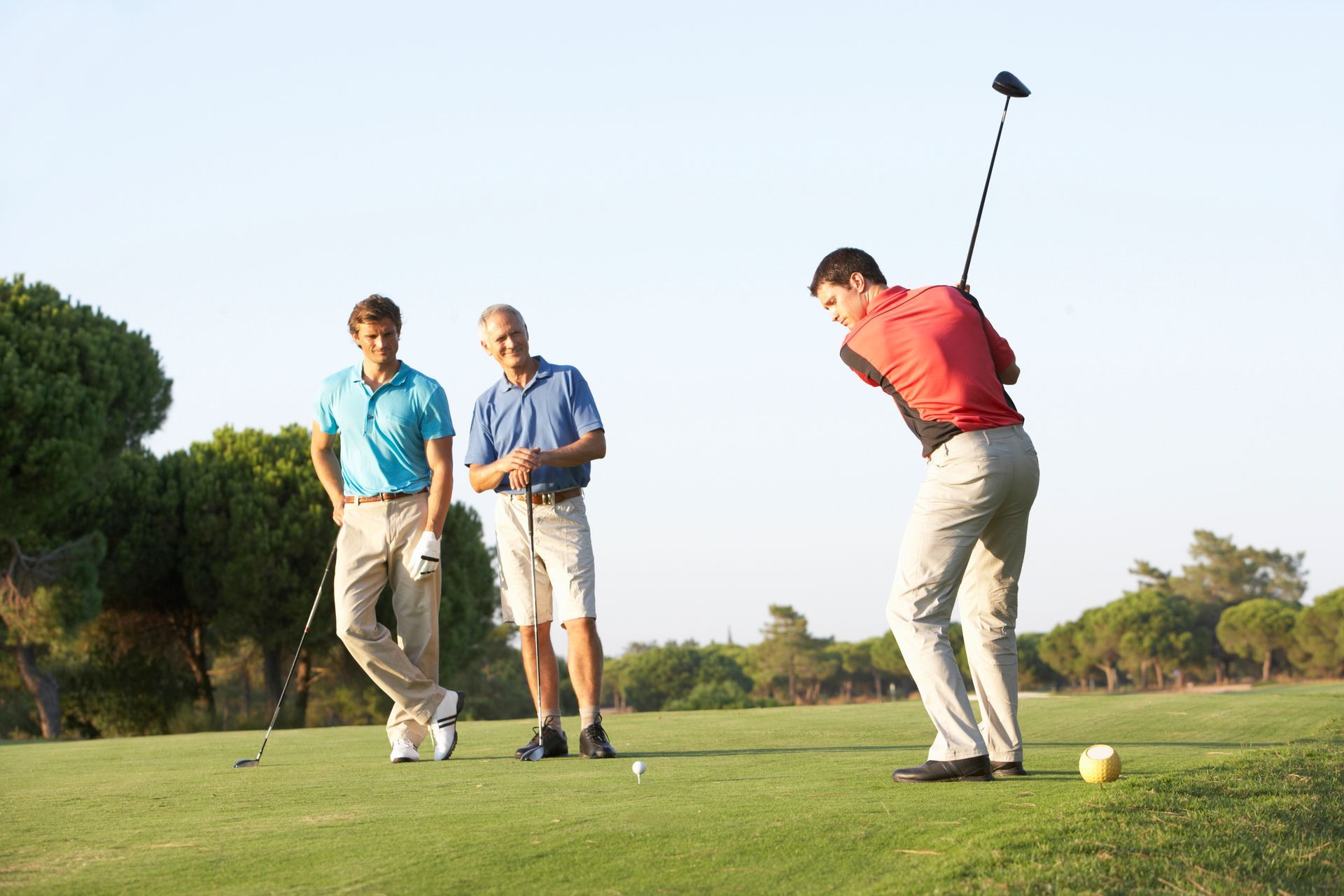 A golfer in a red shirt swings a club on a grassy golf course while two other people watch from the sidelines.
