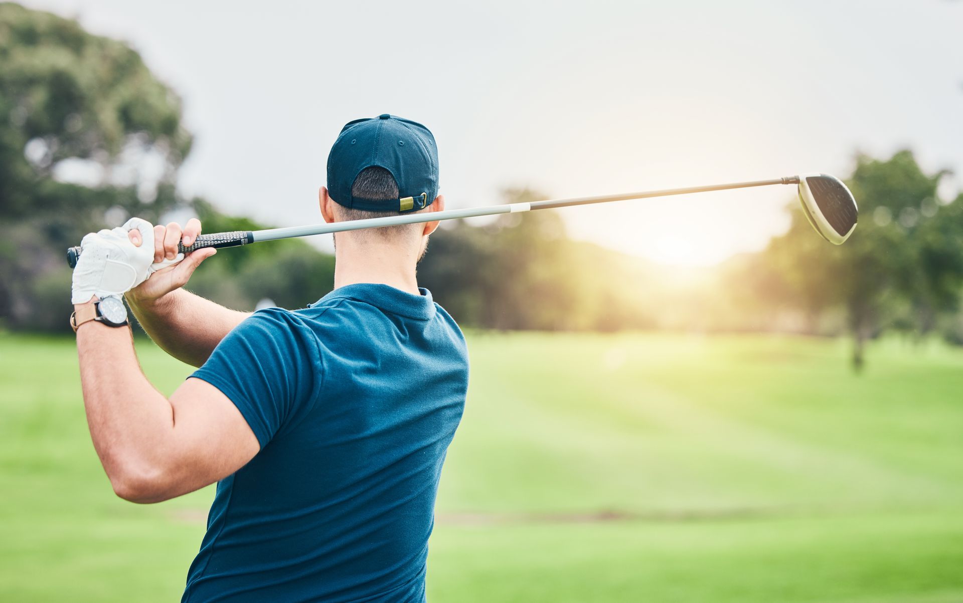 A person wearing a blue shirt and a blue cap is swinging a golf club on a sunlit golf course; trees can be seen in the background.