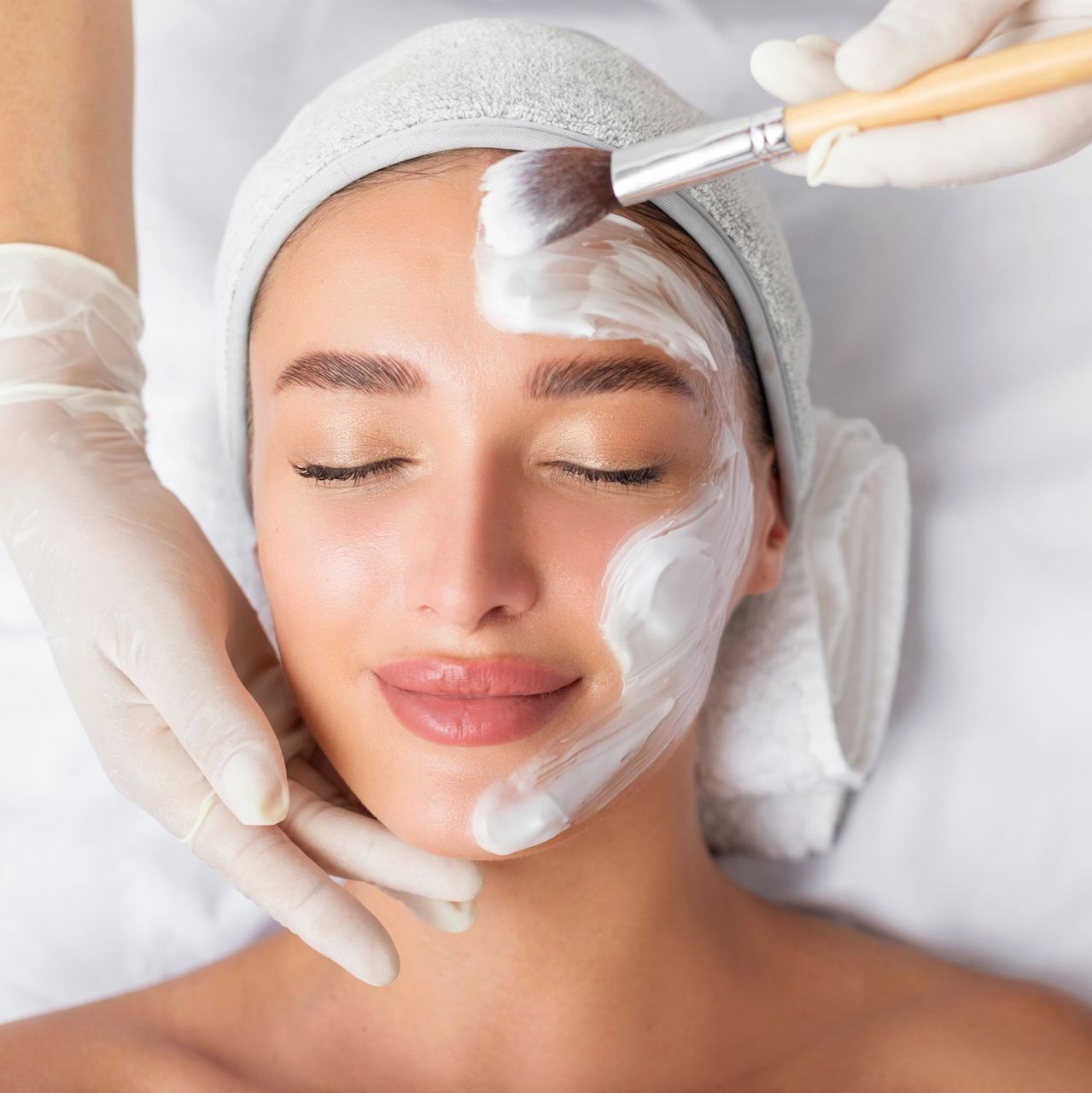 A person with their eyes closed is receiving a facial treatment in which white cream is applied to their face with gloved hands using a brush.