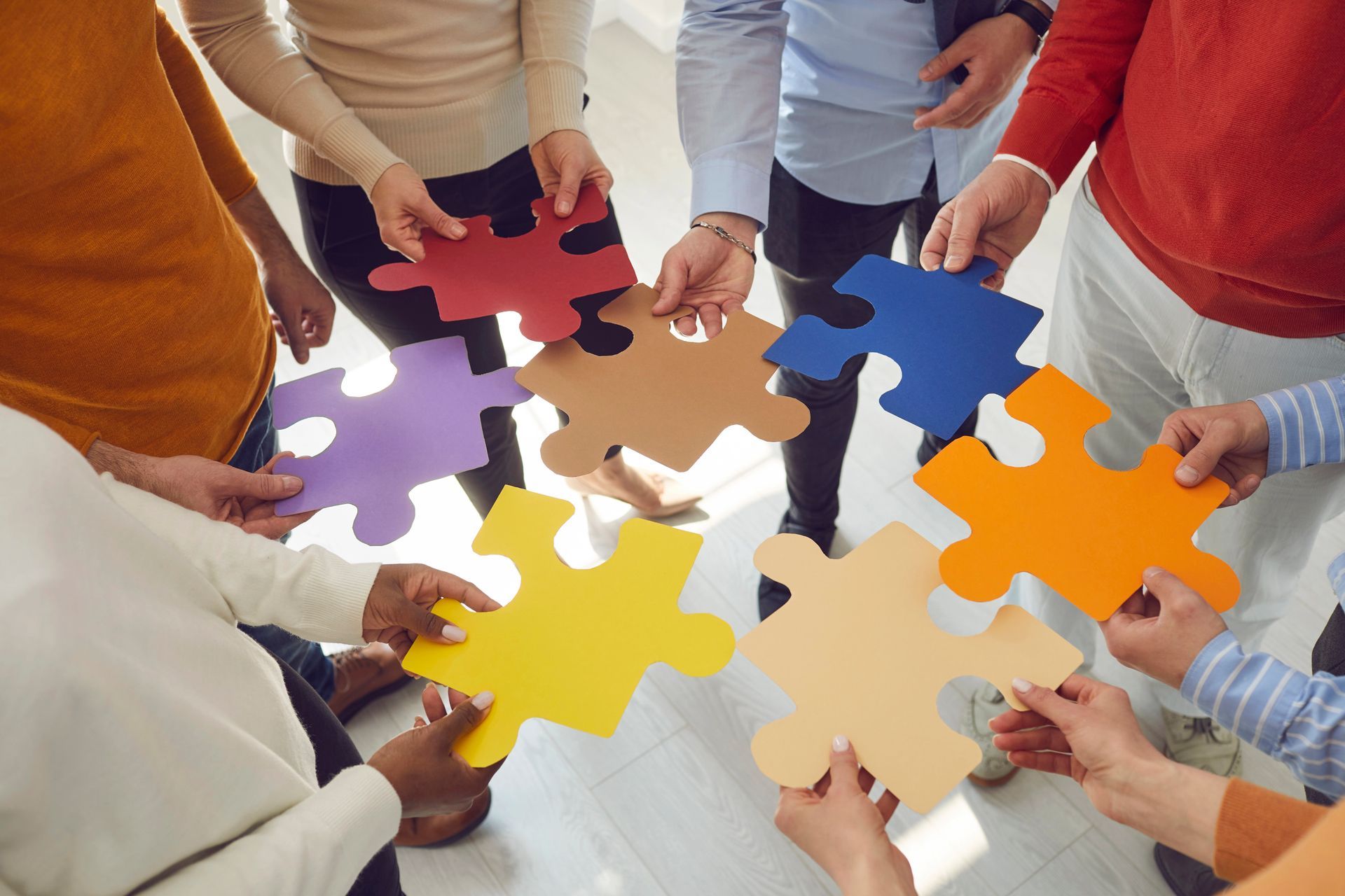 Een groep mensen staat in een kring en houdt puzzelstukjes in verschillende kleuren samen in de richting van het midden.