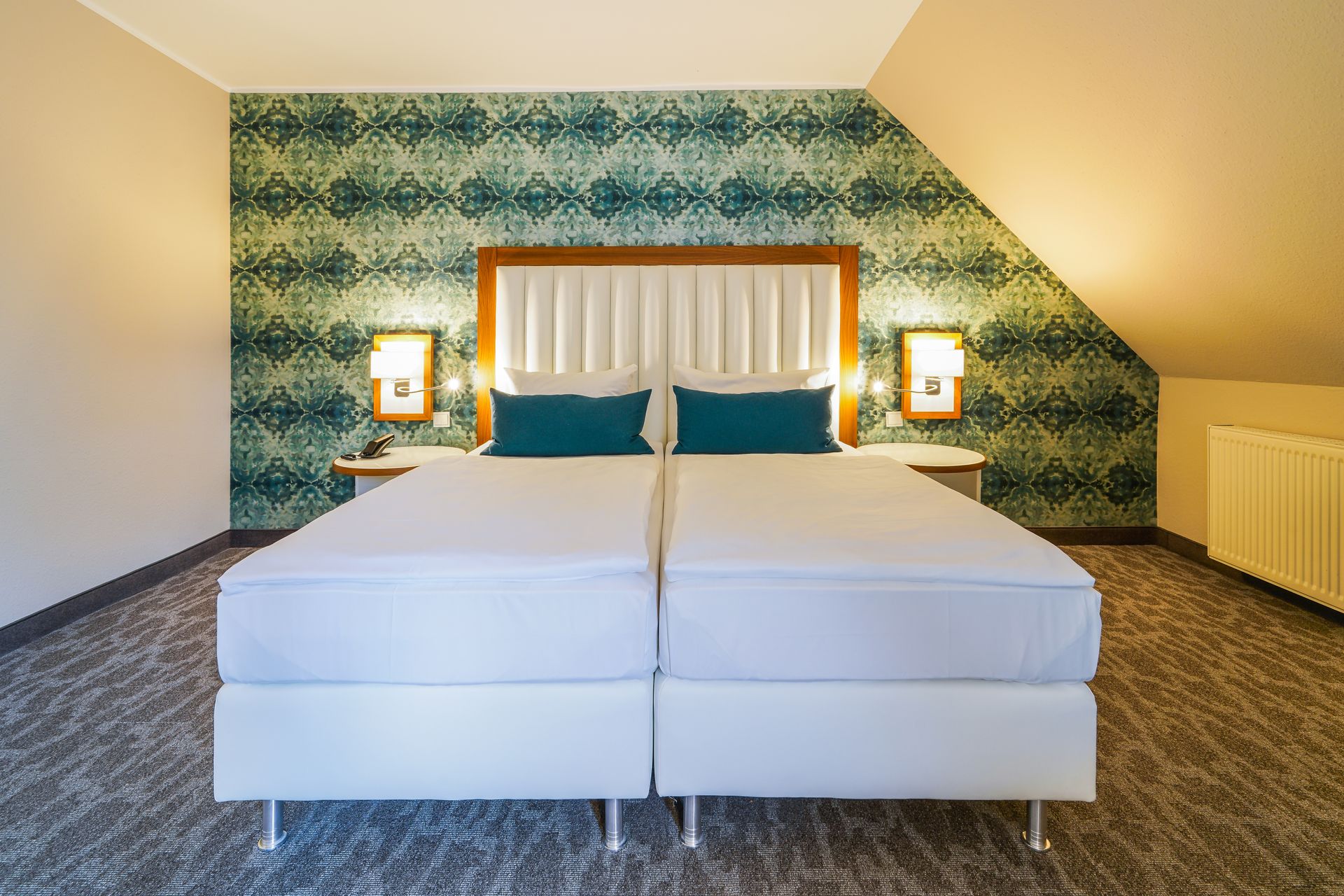 A hotel room with two white twin beds, patterned turquoise wallpaper, and a wooden headboard beneath a sloped ceiling.