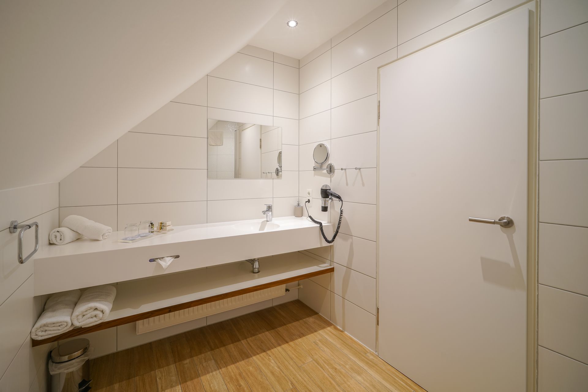 A modern bathroom with a sloped ceiling, a white vanity, a mirror, towels, and a white door on a light-colored wood floor.