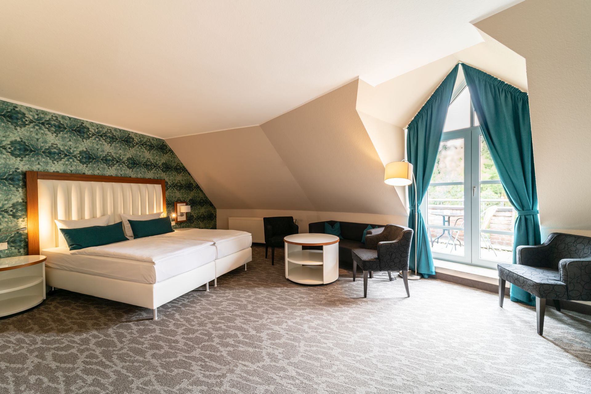 Ein Hotelzimmer mit einem großen Bett, einer gemusterten Tapete, dunklen Akzentstühlen, einem kleinen Tisch und einem Fenster mit türkisfarbenen Vorhängen.