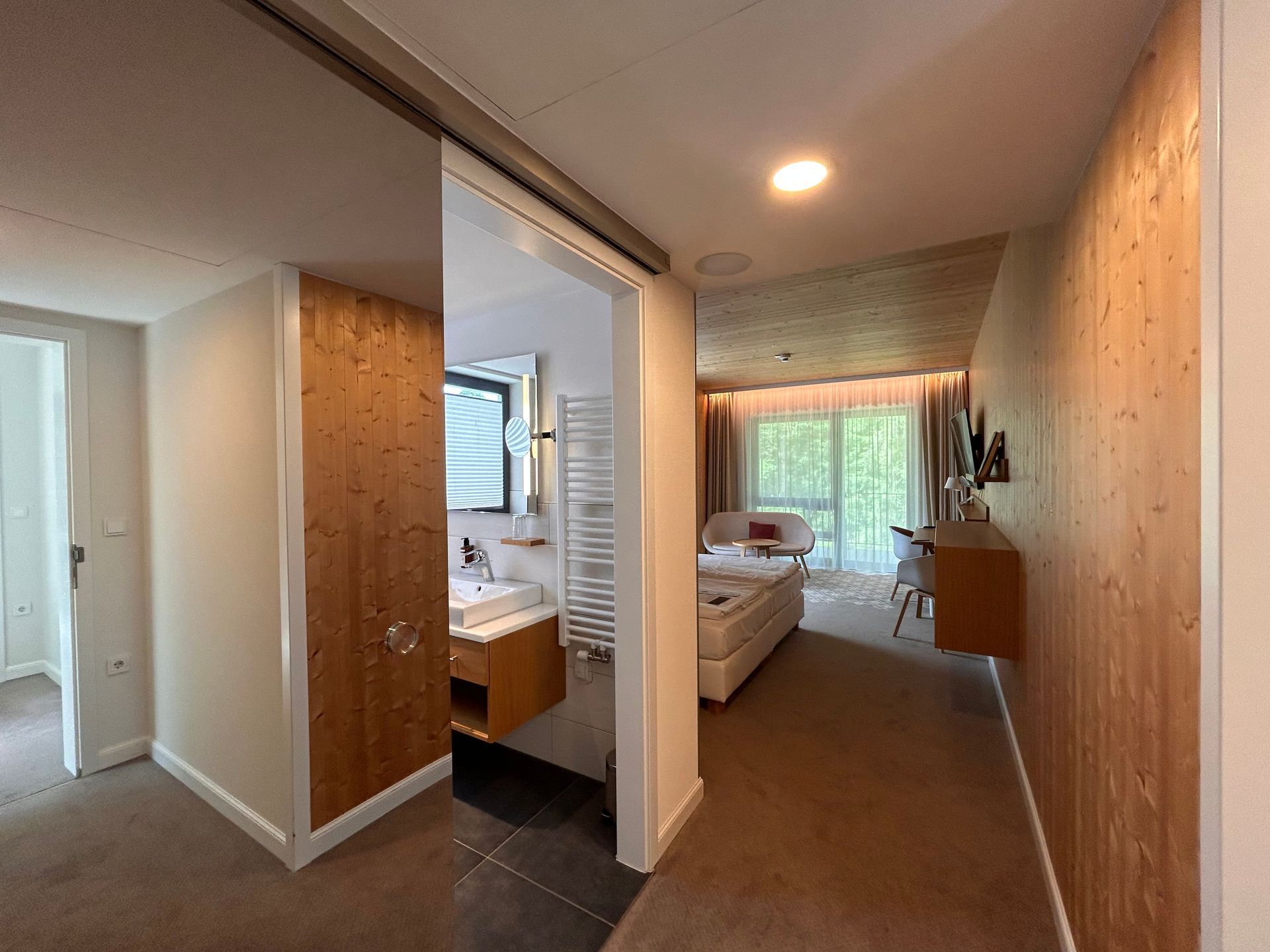 Ein Flur führt vorbei an einem holzgetäfelten Badezimmer in ein helles, mit Teppichboden ausgelegtes Schlafzimmer mit großen Fenstern und weißen Möbeln.