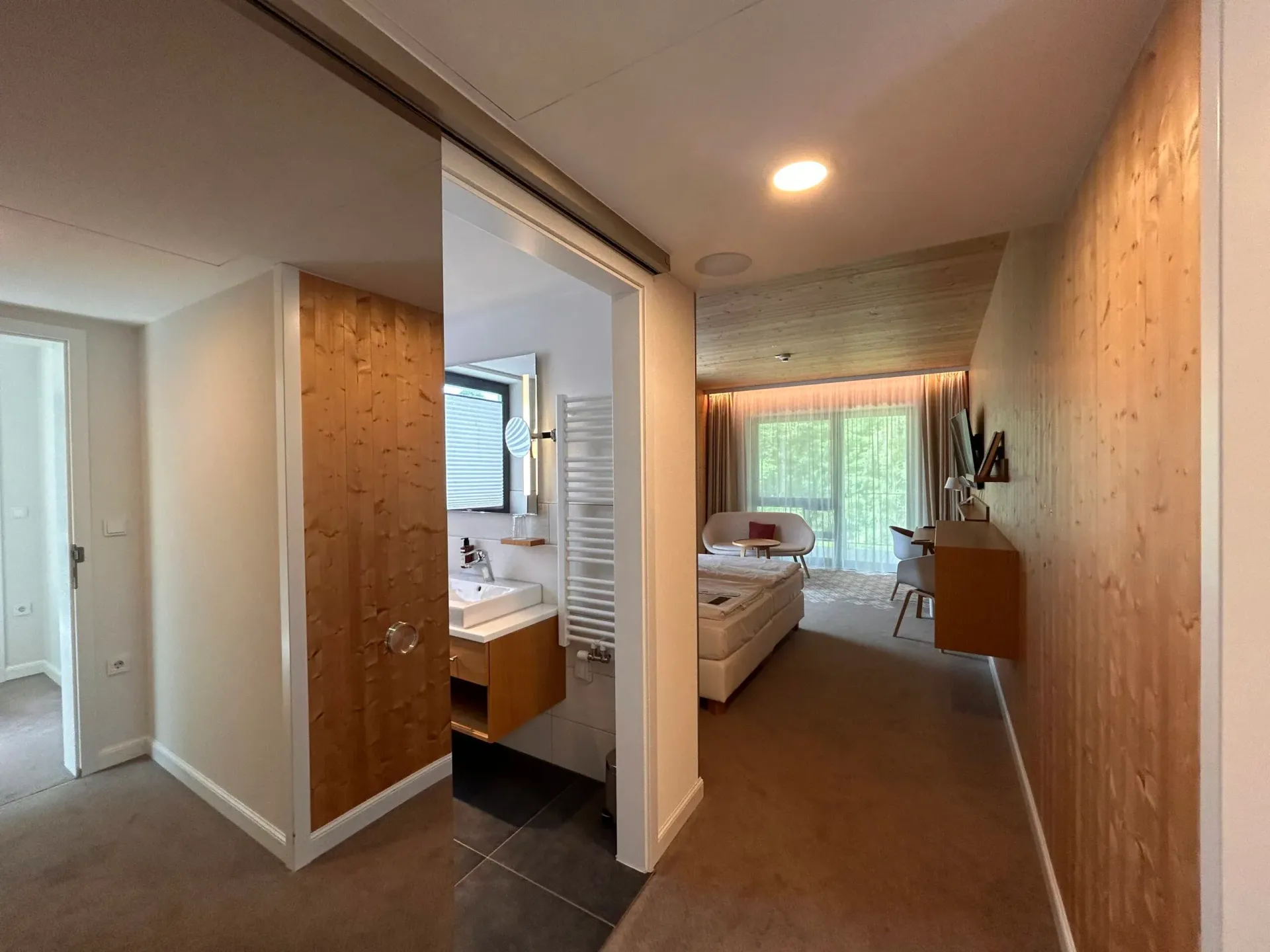 Blick aus einer Hotelsuite mit Flur, holzgetäfeltem Badezimmer und Schlafzimmer mit Bett und bodentiefen Fenstern.