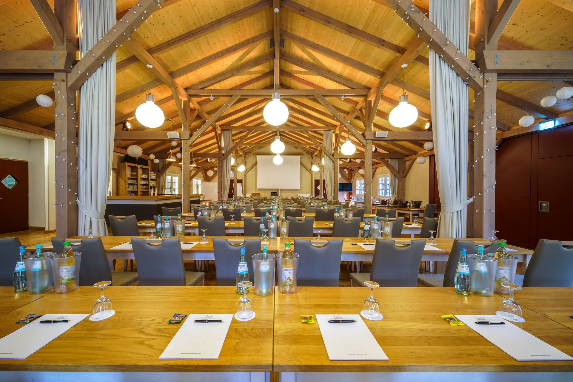 A conference room with long wooden tables, gray chairs, and a vaulted wooden ceiling illuminated by hanging globe lamps.