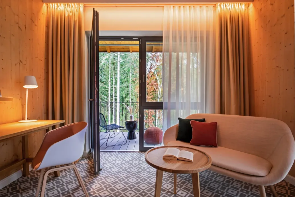 Ein gemütliches Hotelzimmer mit holzgetäfelten Wänden, einem Schreibtisch mit Lampe, einem Sofa mit Zierkissen und Blick auf einen Wald.