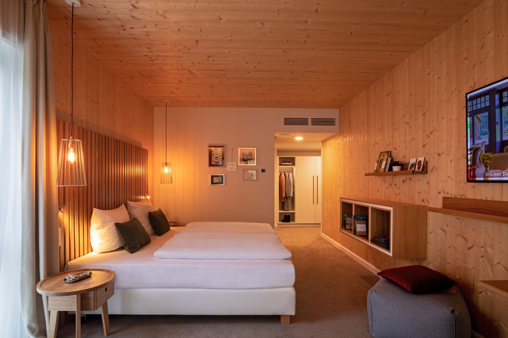 Ein modernes Hotelzimmer mit holzgetäfelten Wänden, einem großen weißen Bett, Nachttischlampen, Staufächern und einem an der Wand montierten Fernseher.