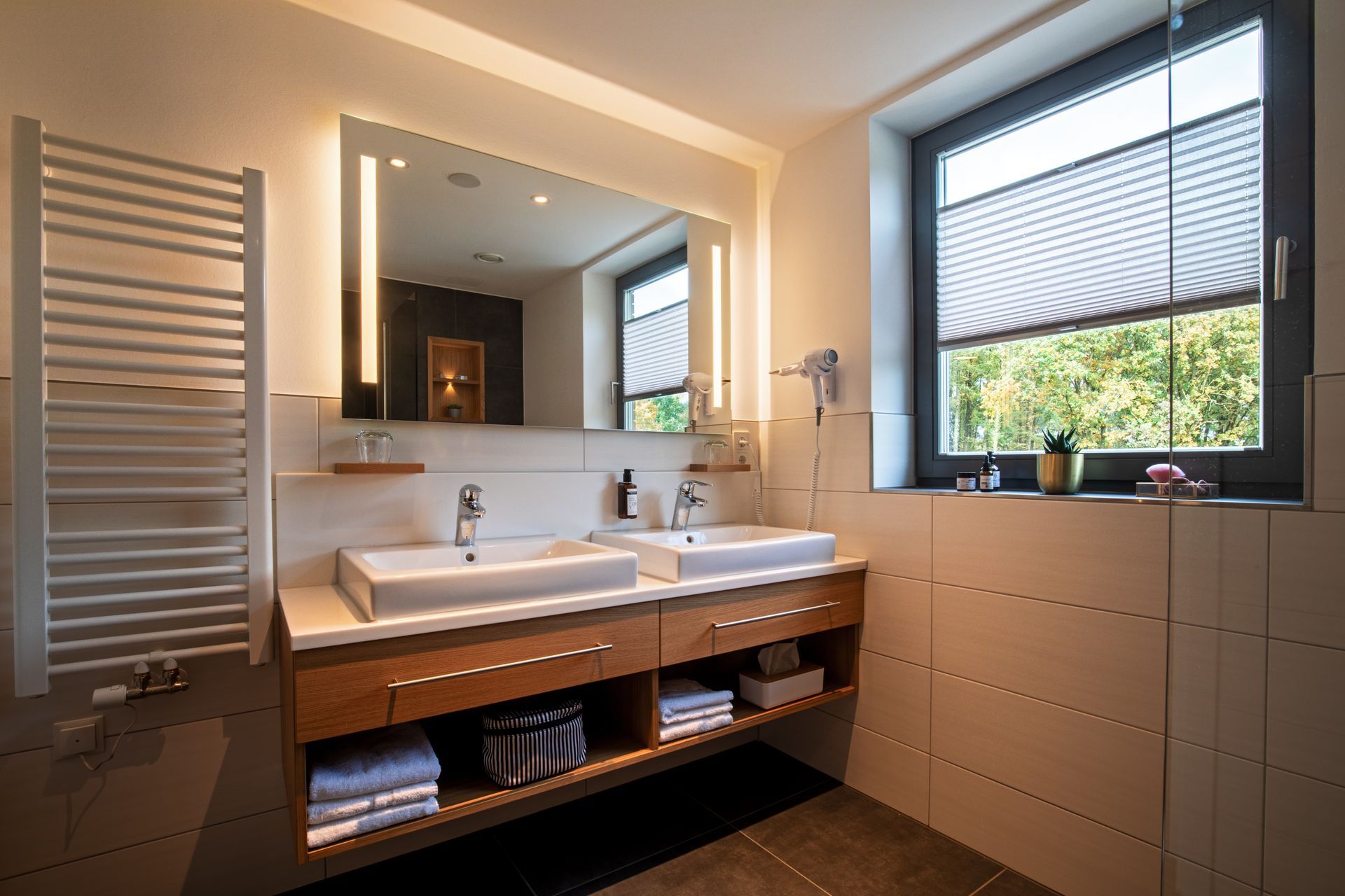 A modern bathroom with a double sink, wooden cabinets with open shelves, a large lighted mirror, and a window.