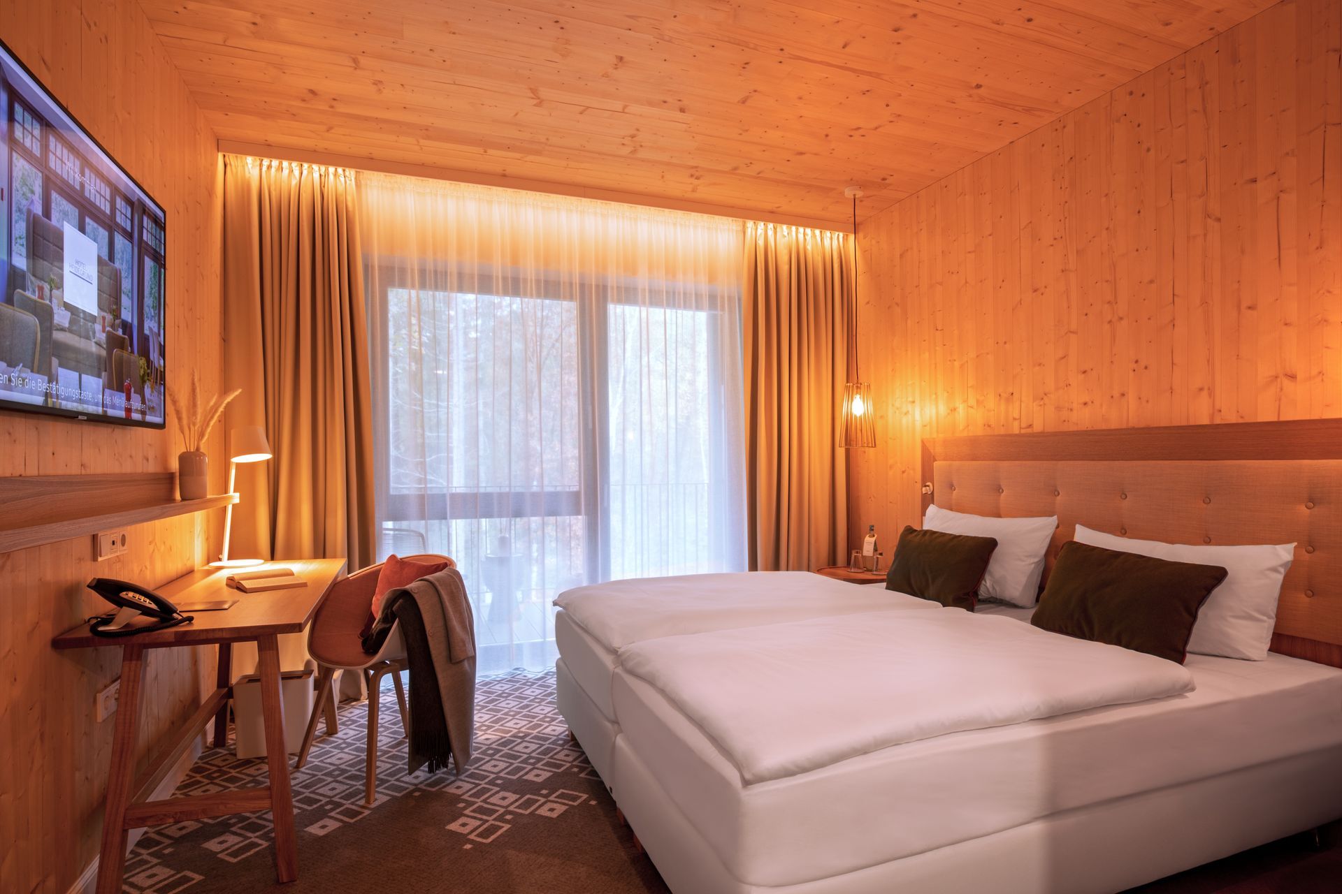 A modern hotel room with wood-paneled walls, a double bed with dark pillows, a wooden desk, and a large window with curtains.