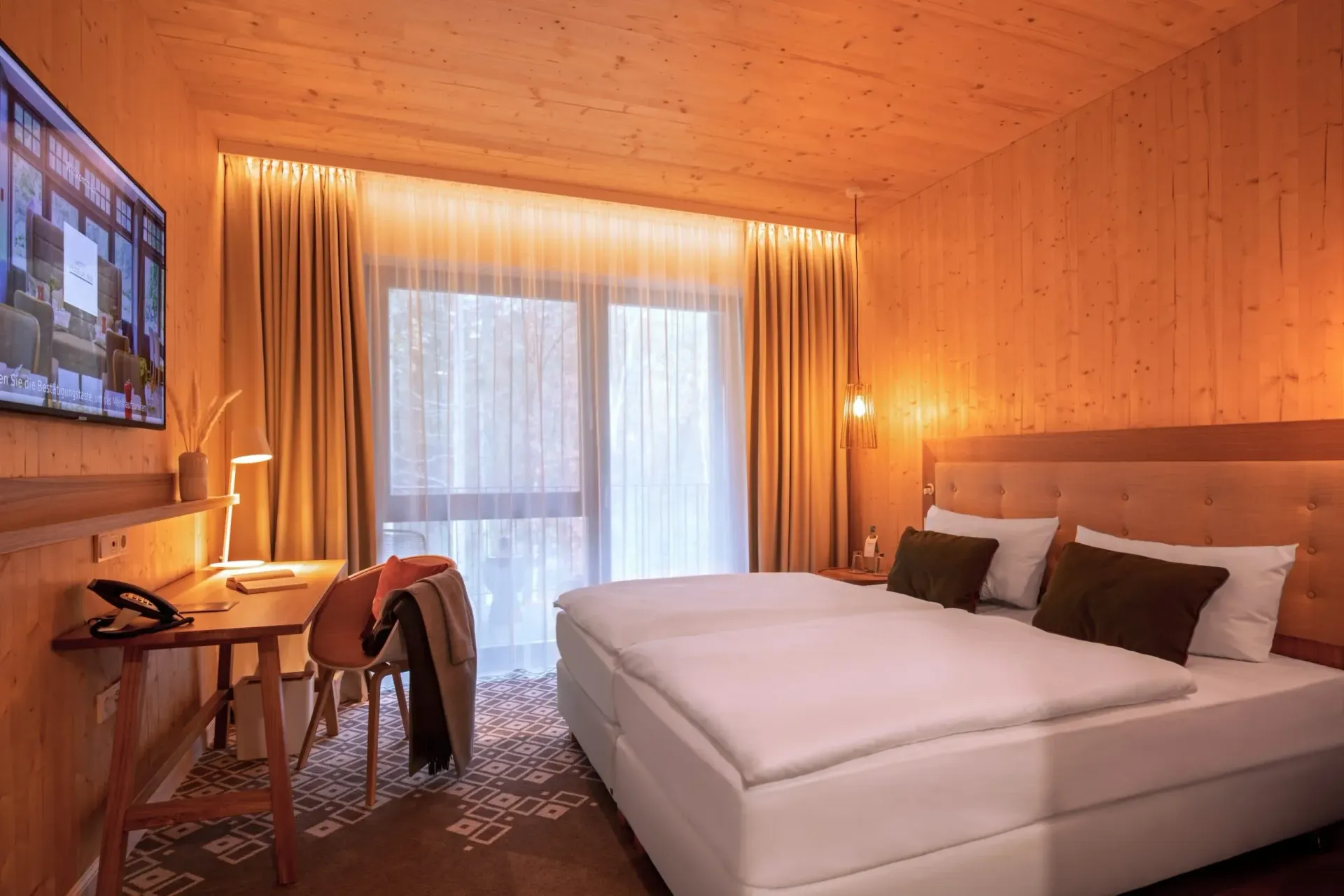A hotel room with a queen-size bed, floral wallpaper, a desk, and a window with curtains overlooking a terrace.