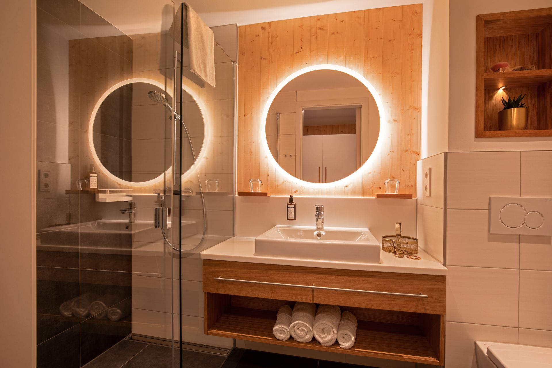 Modernes Badezimmer mit holzgetäfelter Wand, beleuchtetem runden Spiegel, weißem Aufsatzwaschbecken und gefalteten Handtüchern.