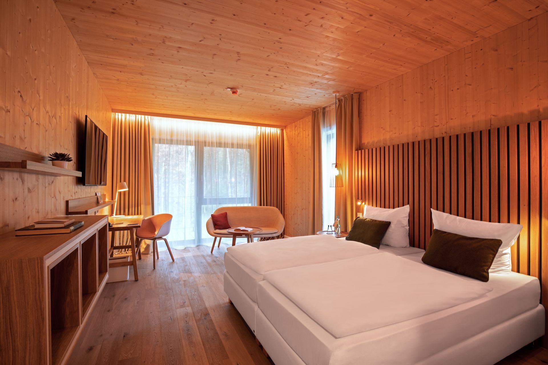 Ein modernes Hotelzimmer mit holzgetäfelten Wänden, einem großen Bett mit dunklen Kissen, einer Sitzecke und einem bodentiefen Fenster.