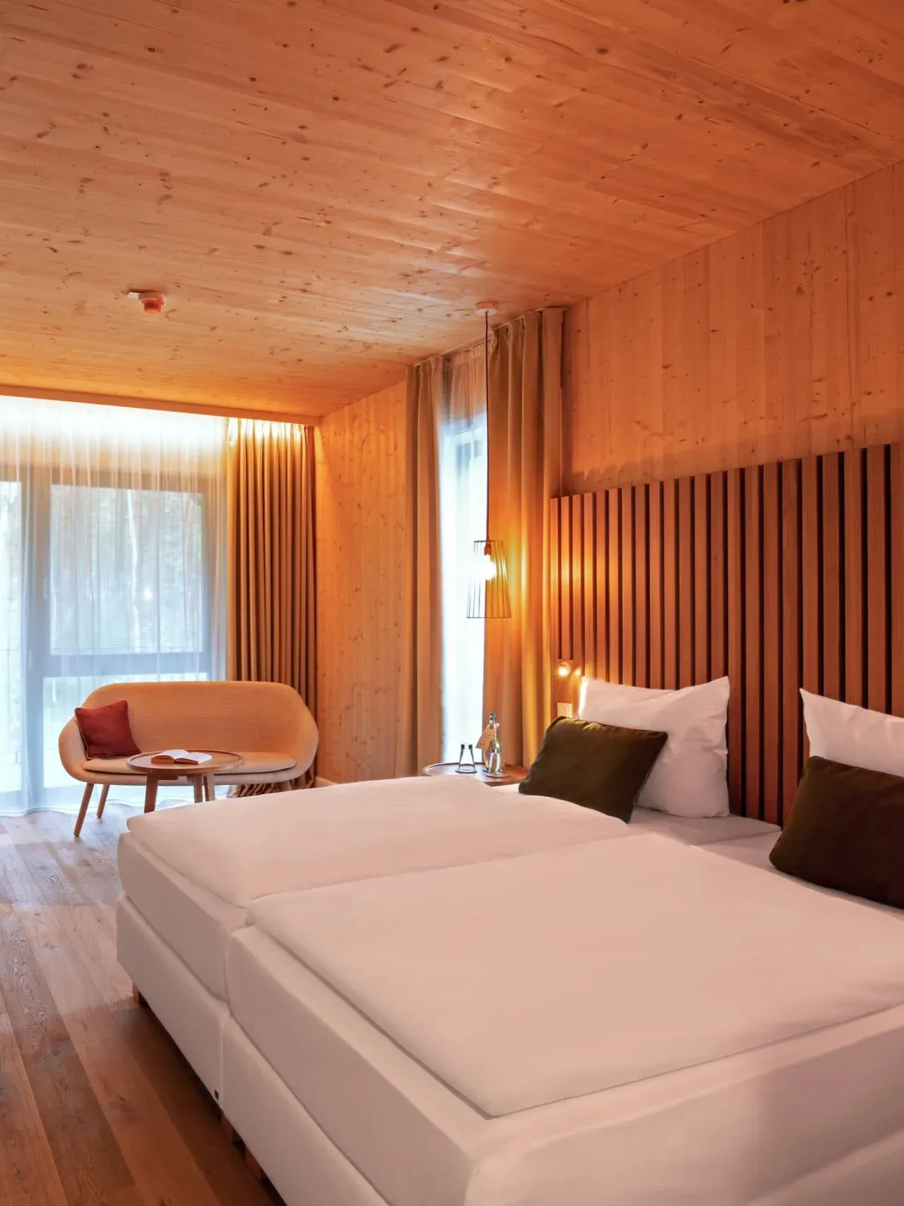A hotel room with wood-paneled walls and ceiling, a large bed with white linens, and a small sofa by a bright window.