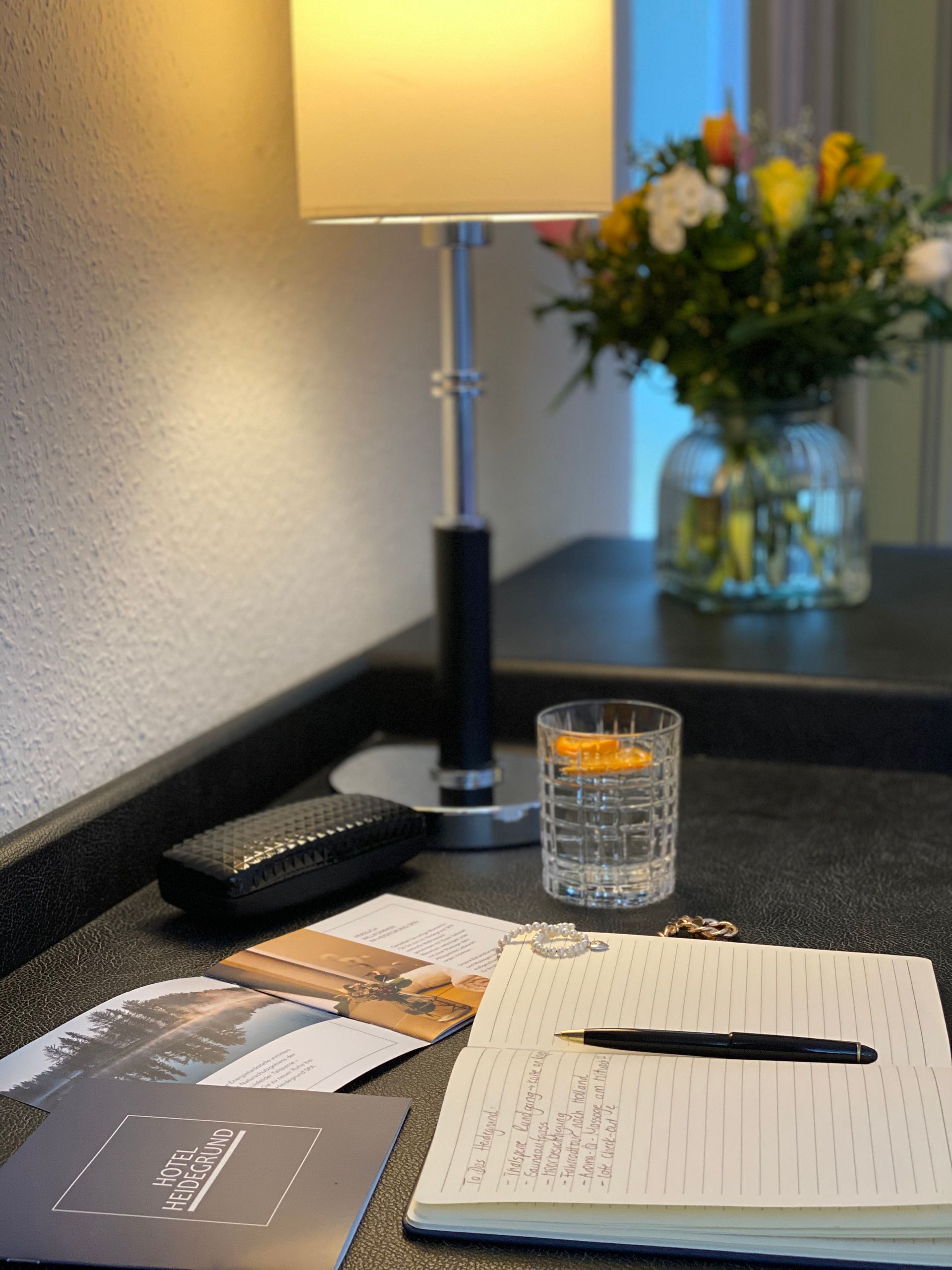 Ein Tisch mit einer Lampe, einem Glas Wasser, einem Blumenstrauß und einem Notizbuch mit Stift auf einer dunklen Oberfläche.
