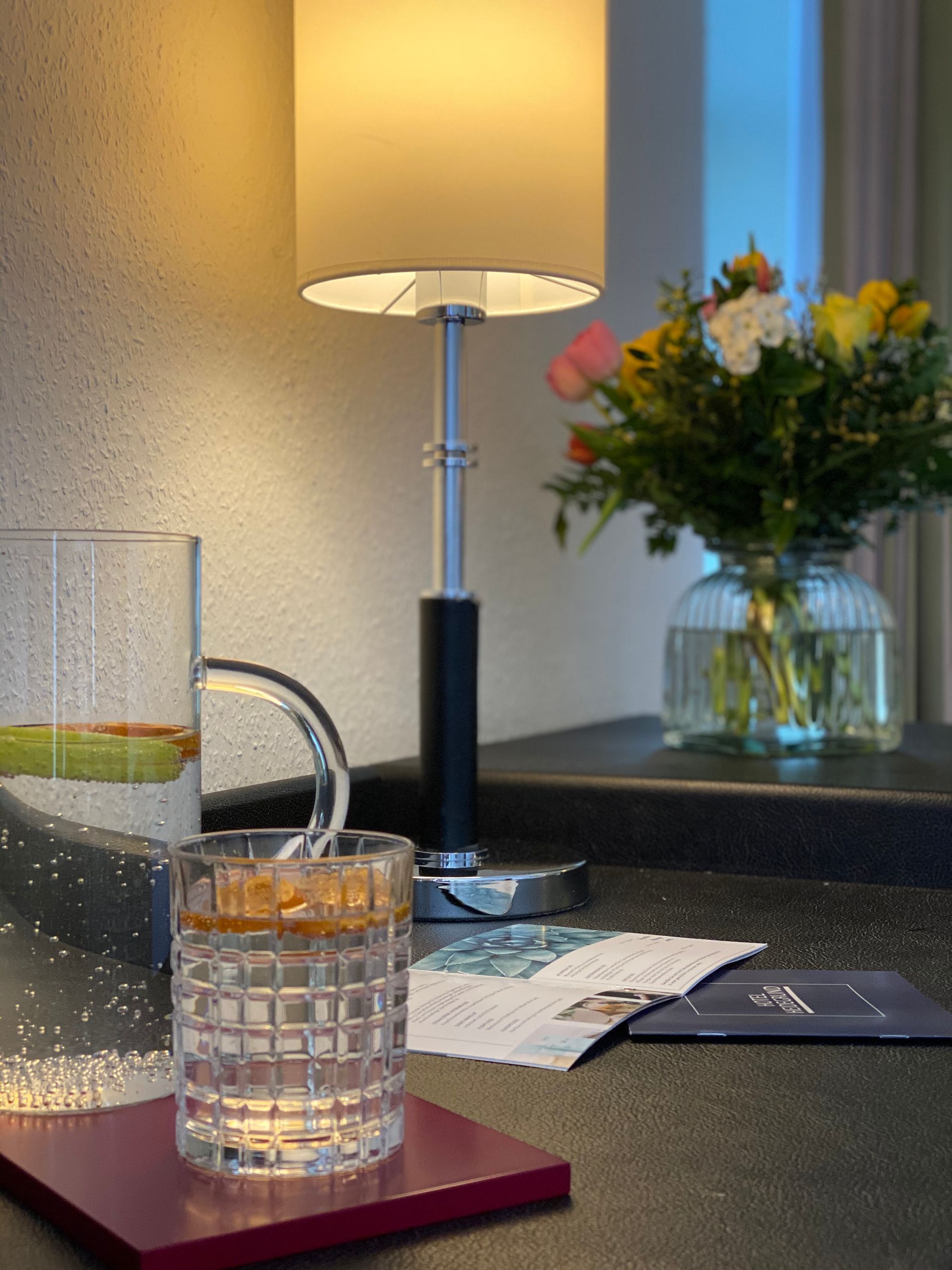 Op een donkere bar staat naast een glas water een moderne lamp; op de achtergrond is een vaas met bloemen te zien.