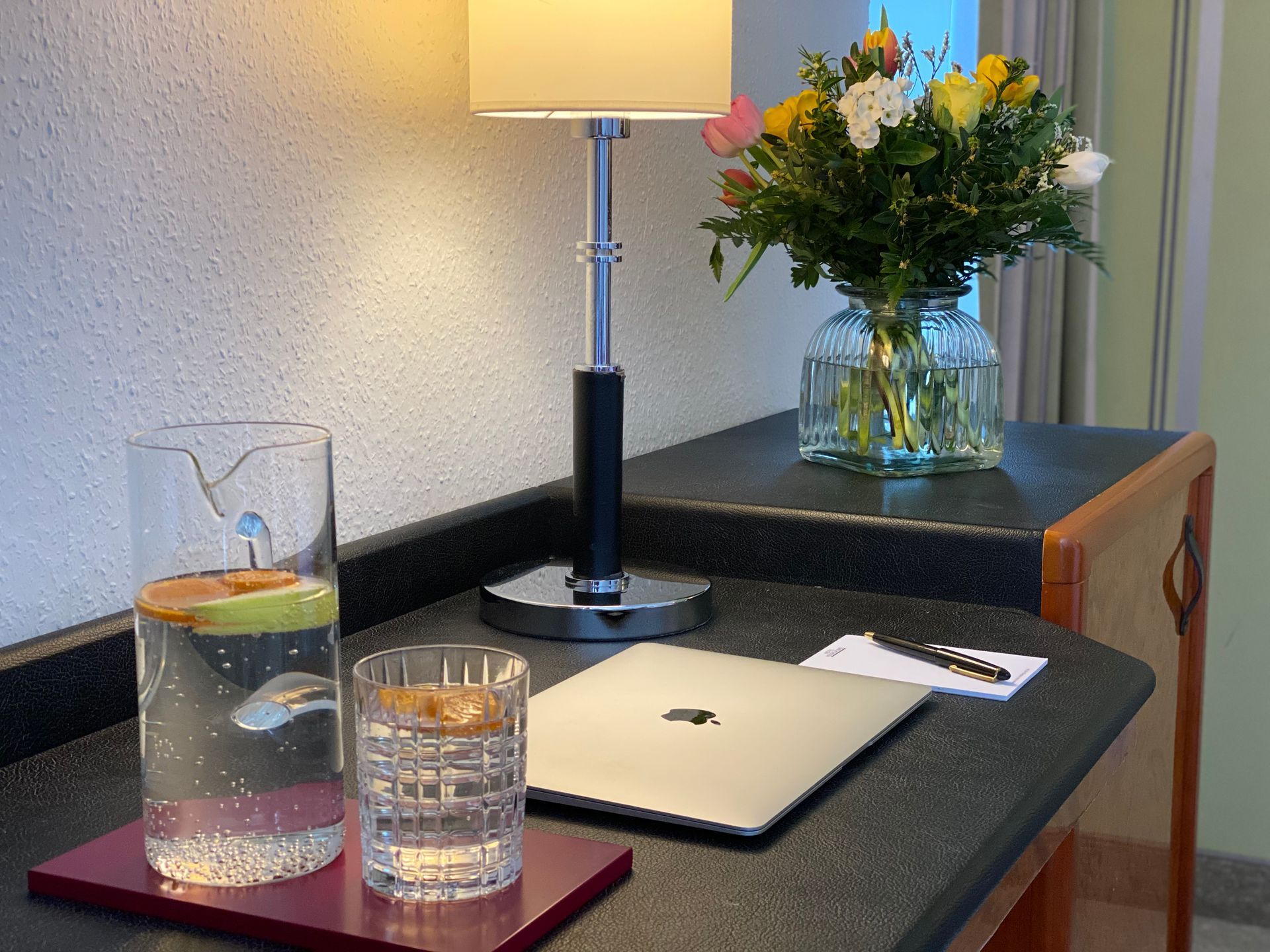 Ein schwarzes Kleid, eine Perlenkette, ein Goldring und eine schwarze Clutch liegen auf einem Bett mit bunten Zierkissen.