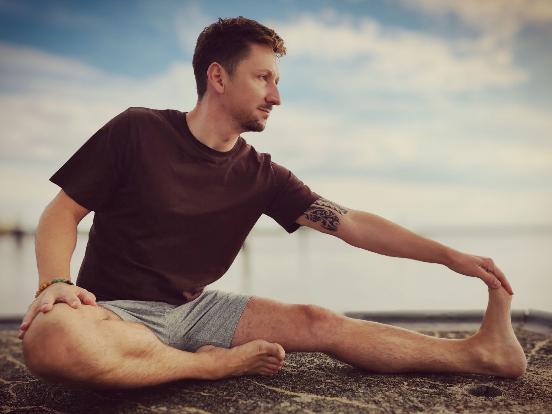 Eine Person, die ein braunes T-Shirt und graue Shorts trägt, führt im Sitzen auf einem Pier am Wasser eine Dehnung der hinteren Oberschenkelmuskulatur durch.