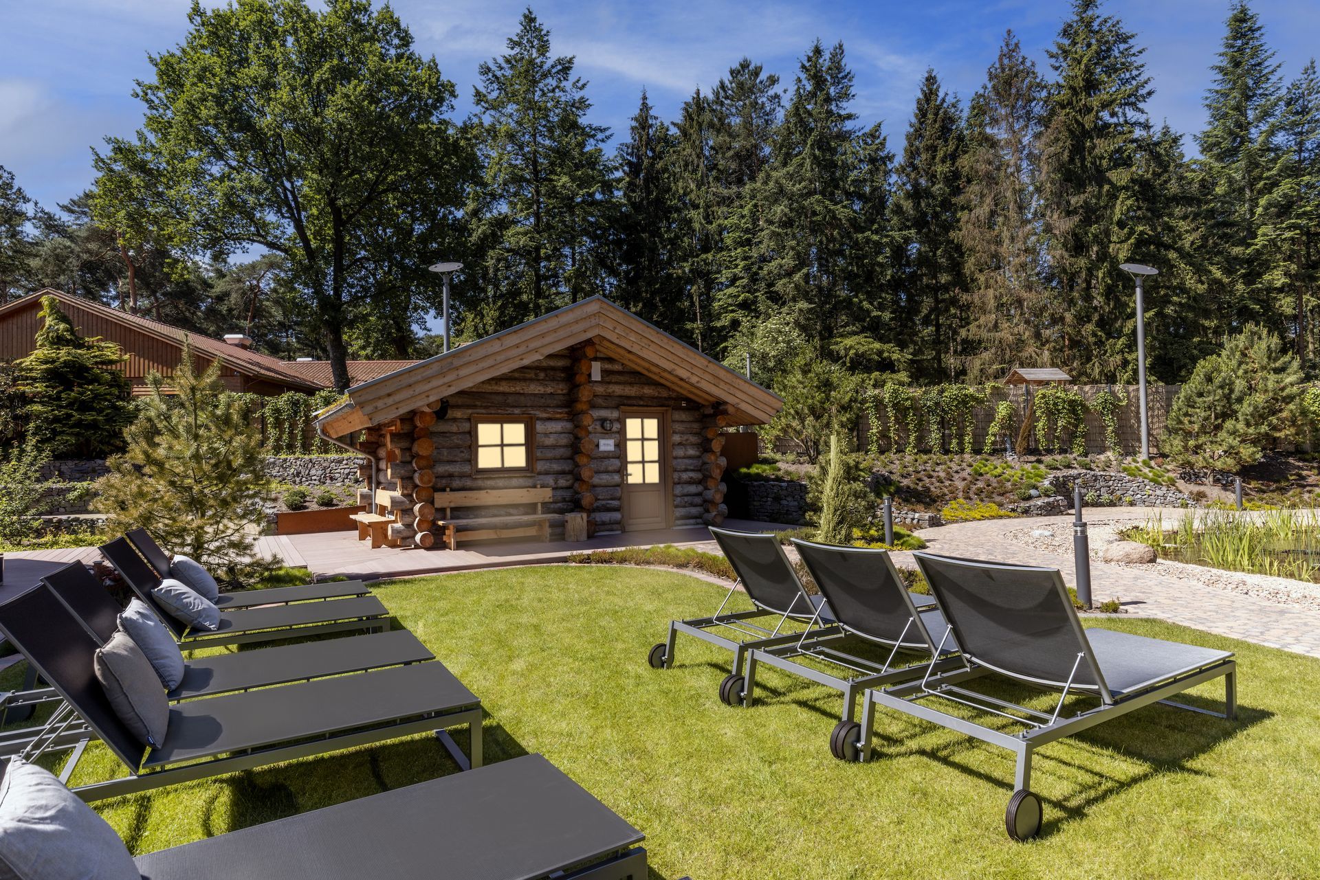 Auf einer Rasenfläche vor einer rustikalen Blockhütte unter strahlend blauem Himmel steht eine Reihe von Liegestühlen.
