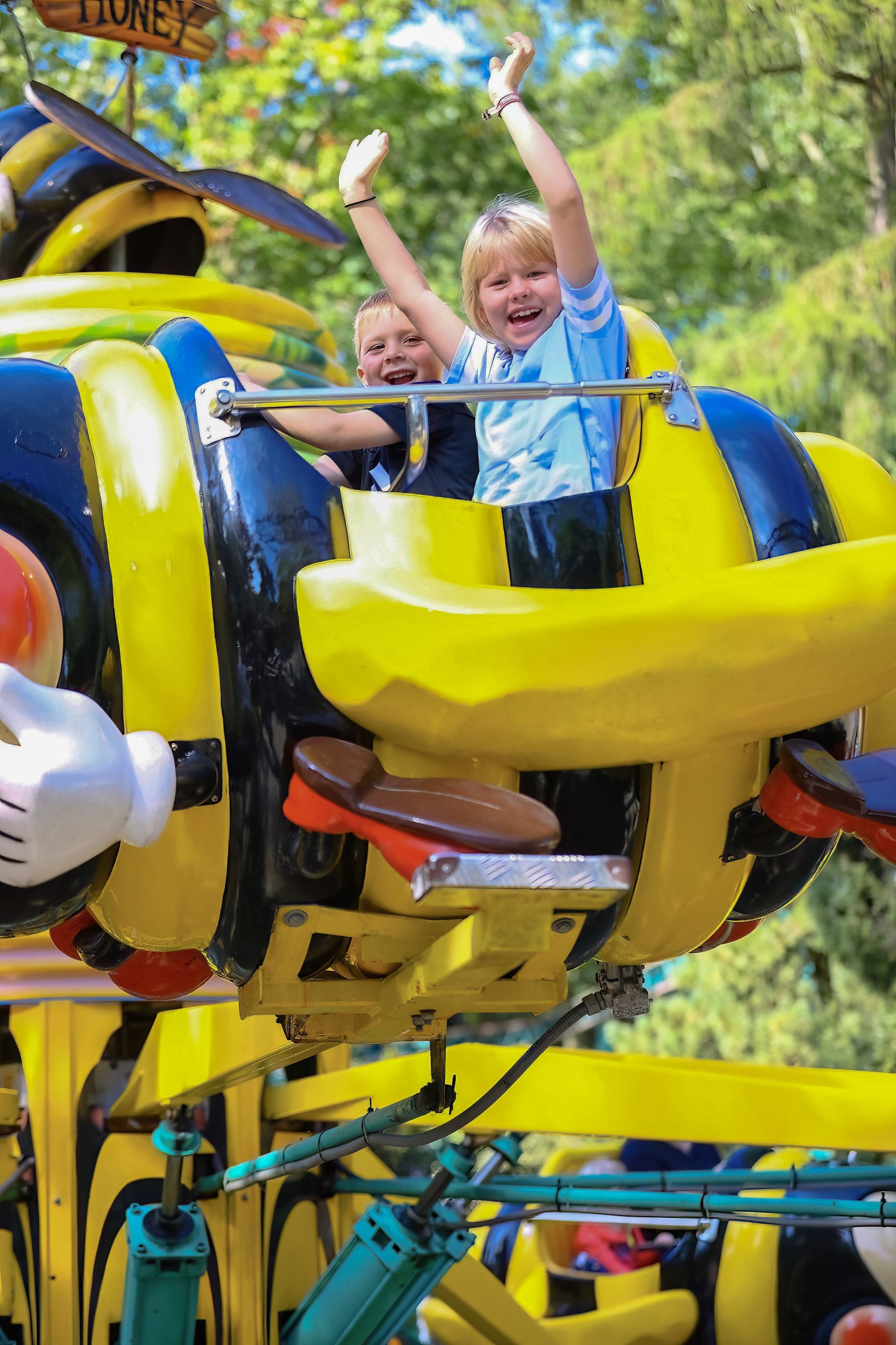 Two children are smiling with their arms raised on a yellow, bee-shaped carousel.