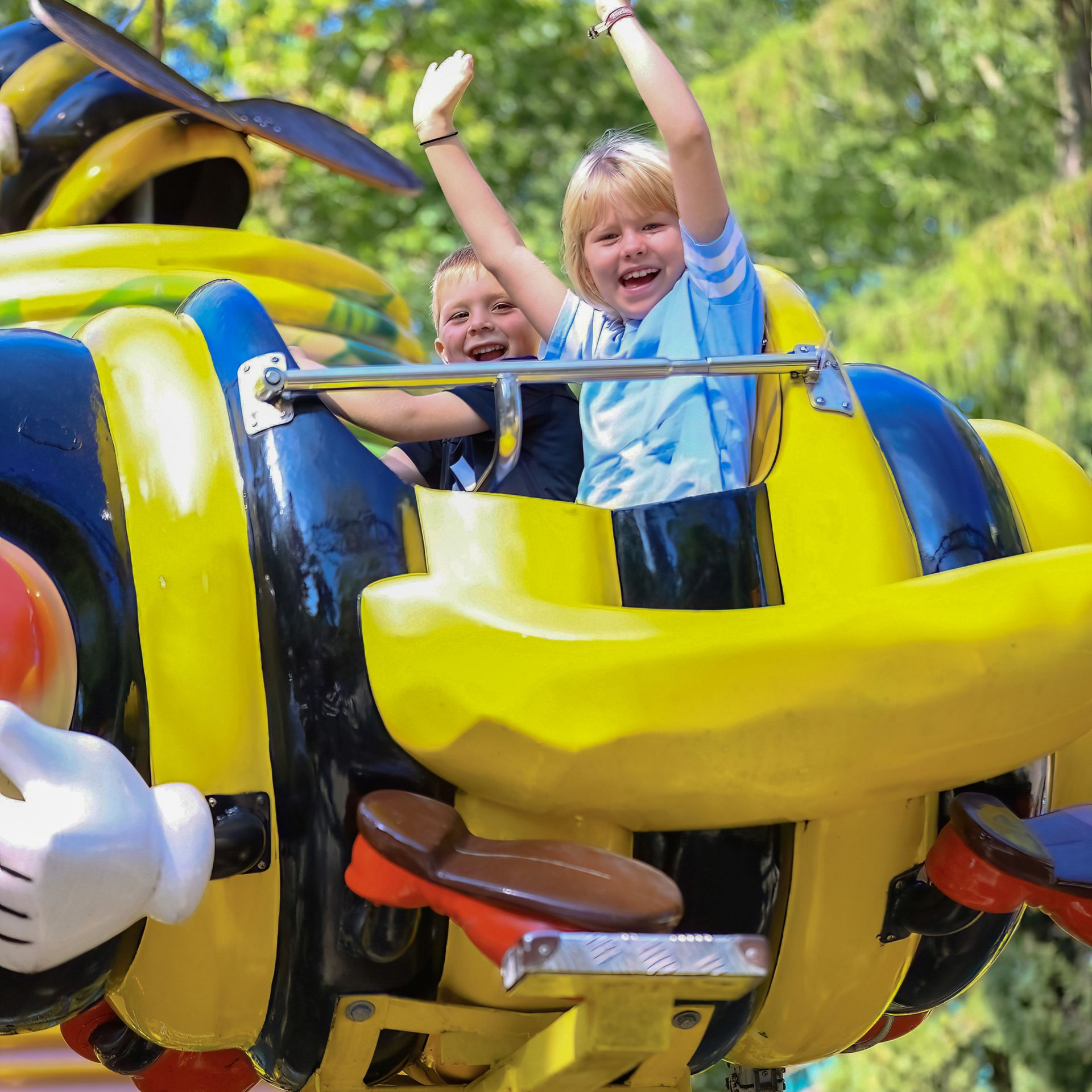 Two children with excited expressions are riding on a yellow-and-blue bee-shaped ride, their hands raised high.